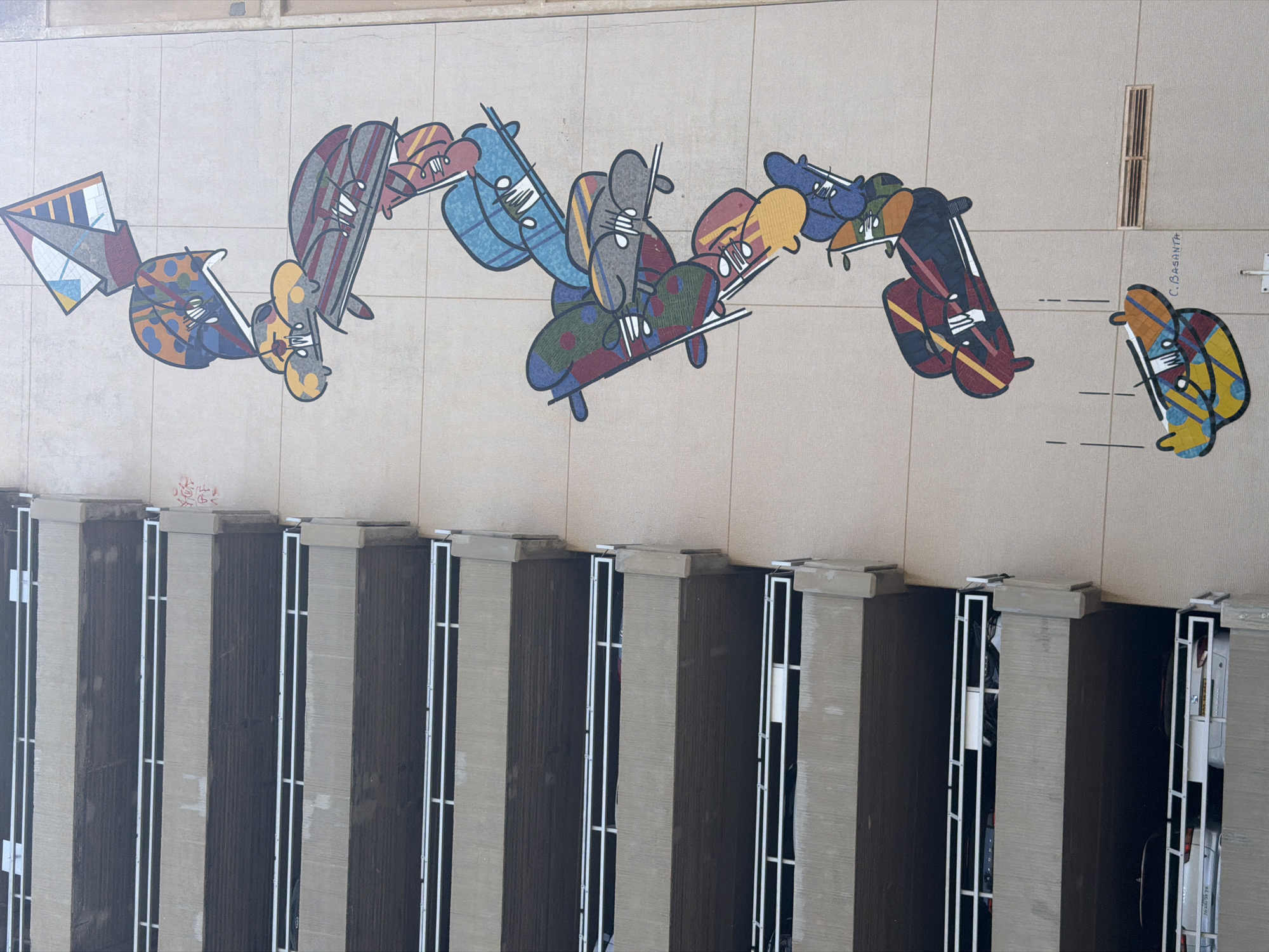 The image shows an aerial view of a mural and a building in Tel Aviv, Israel. The mural, located on a light beige wall, features a colorful design of interconnected abstract figures, resembling people on skateboards. The artist's signature, "C. BASANTA", is visible next to the last figure. Below the mural, the top of a building is visible, consisting of a row of evenly spaced, gray columns. Each column has a square concrete cap. The spaces between the columns are narrow and enclosed by white metal railings. Within these spaces, glimpses of interiors, possibly parking garages, are visible, including cars and other objects. The overall composition suggests an urban environment where art is integrated into the architectural landscape.
