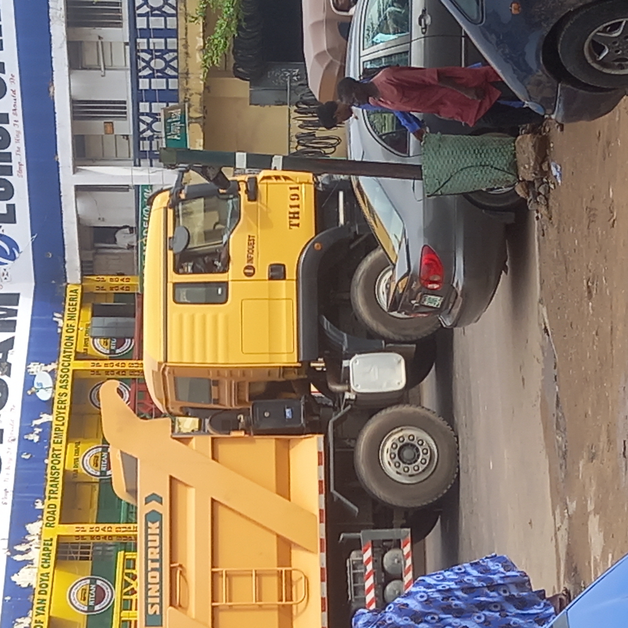 The image shows a yellow Sinotruk tipper truck on a street in Sardauna, Nigeria. The truck has "INFIQUEST TH191" printed on its side. Next to the truck is a gray car and a person wearing a red shirt. The background includes a sign for "OF YAN DOYA CHAPEL ROAD TRANSPORT EMPLOYER'S ASSOCIATION OF NIGERIA." A blue and white patterned cloth is partially visible at the bottom of the frame.