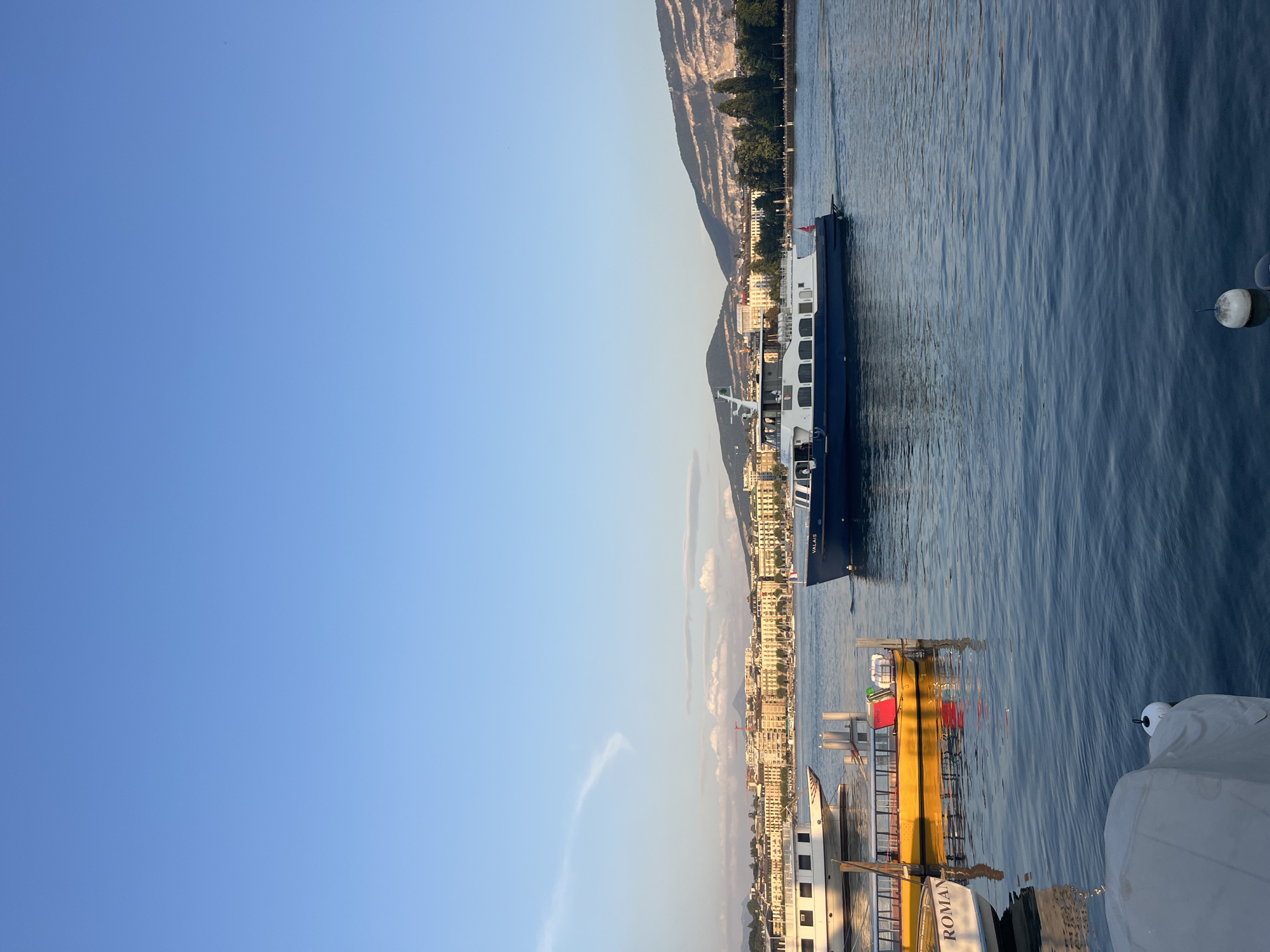 This image depicts a view of Lake Geneva in Geneva, Switzerland, during daylight hours. The water is calm and reflects the clear blue sky. In the foreground, several boats are docked. A large white boat with the name "ROMAN" partially visible is on the left, and a yellow boat with red and green accents is next to it. Further out in the water, a blue and white passenger boat is underway. The background features the cityscape of Geneva with numerous buildings and a backdrop of green hills and mountains. The sky is a gradient of light blue, with a wispy cloud formation visible on the left. The overall impression is a serene and picturesque scene of a European city on a lake.