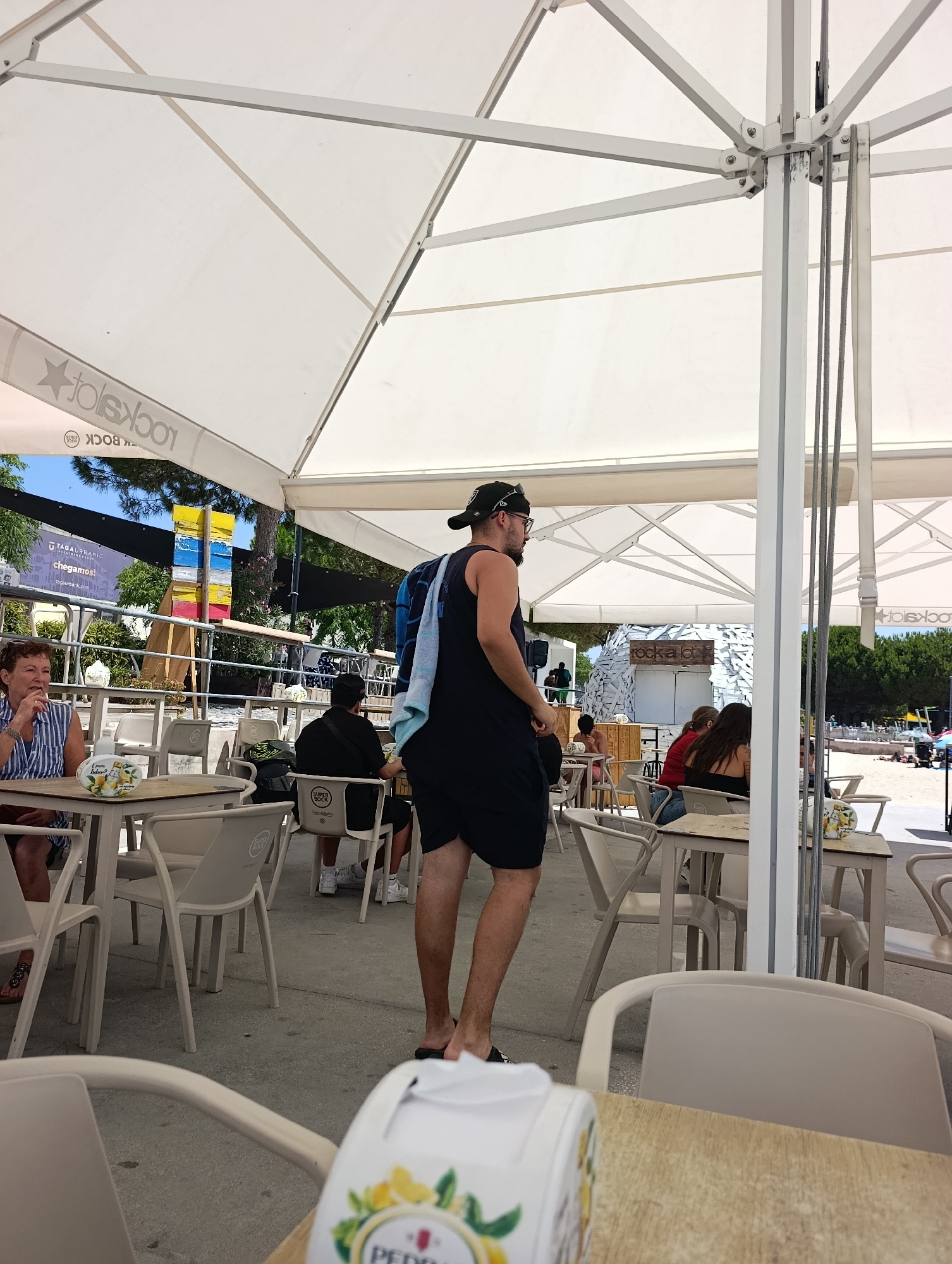 This is an outdoor scene at a beachfront establishment in Setúbal, Portugal. A man wearing a black tank top, black shorts, a black baseball cap, and sunglasses is standing in the foreground, facing away from the camera. He has a blue and white towel draped over his left shoulder. In the background, several people are seated at tables under a white canopy. A woman with curly brown hair wearing a blue and white striped shirt is seated at a table to the left. The establishment appears to be a cafe or restaurant on a beach, with tables and chairs set up on a concrete floor. In the far background, a beach with people can be seen. The overall ambiance is relaxed and casual, consistent with a seaside resort.
