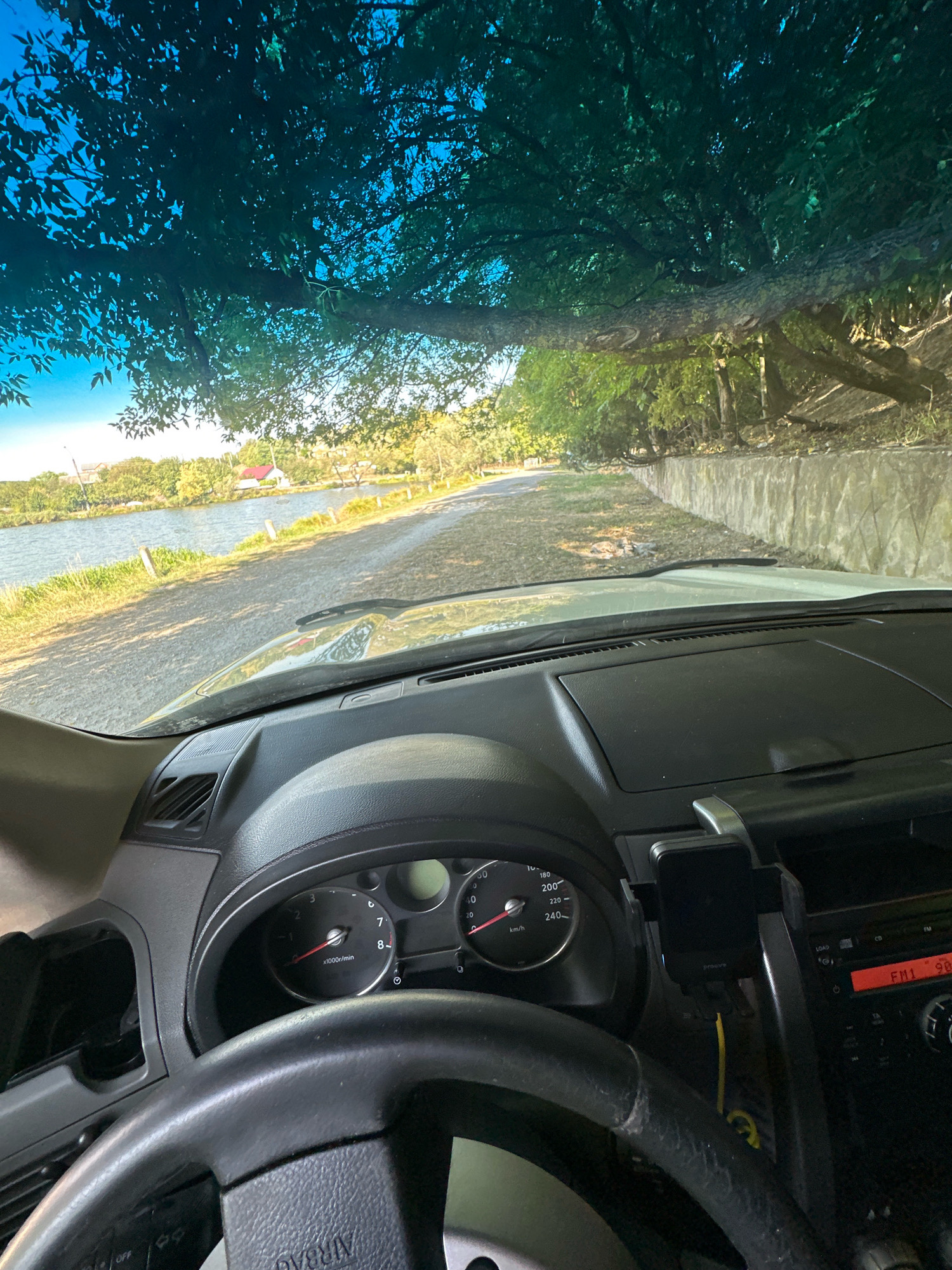 The image is taken from inside a vehicle, looking out through the windshield. The view shows a rural road lined with trees and a body of water on the left. A large tree branches over the windshield, casting shadows. The dashboard and steering wheel of the car are visible in the foreground. The setting appears to be a sunny day with clear blue skies. The location is identified as Makiv, Ukraine.