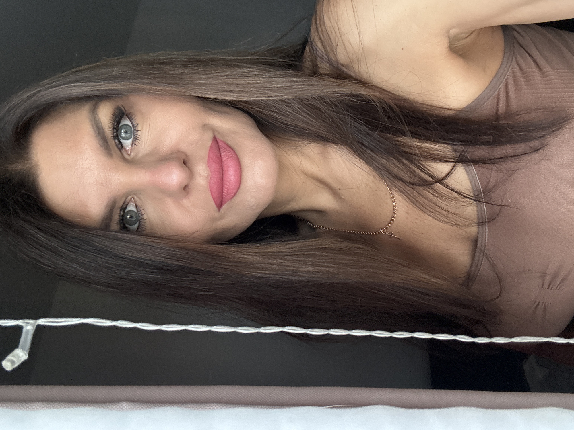 The image shows a close-up, head-and-shoulders portrait of a woman with long, dark brown hair and blue eyes. She is wearing pink lipstick and a brown tank top. A necklace with a cross pendant is visible around her neck. To the left of the woman is a sheer white curtain and a dark background. A string of white lights runs vertically along the left side of the frame. The setting does not provide any specific clues to its location within Chaikovsky, Russia.