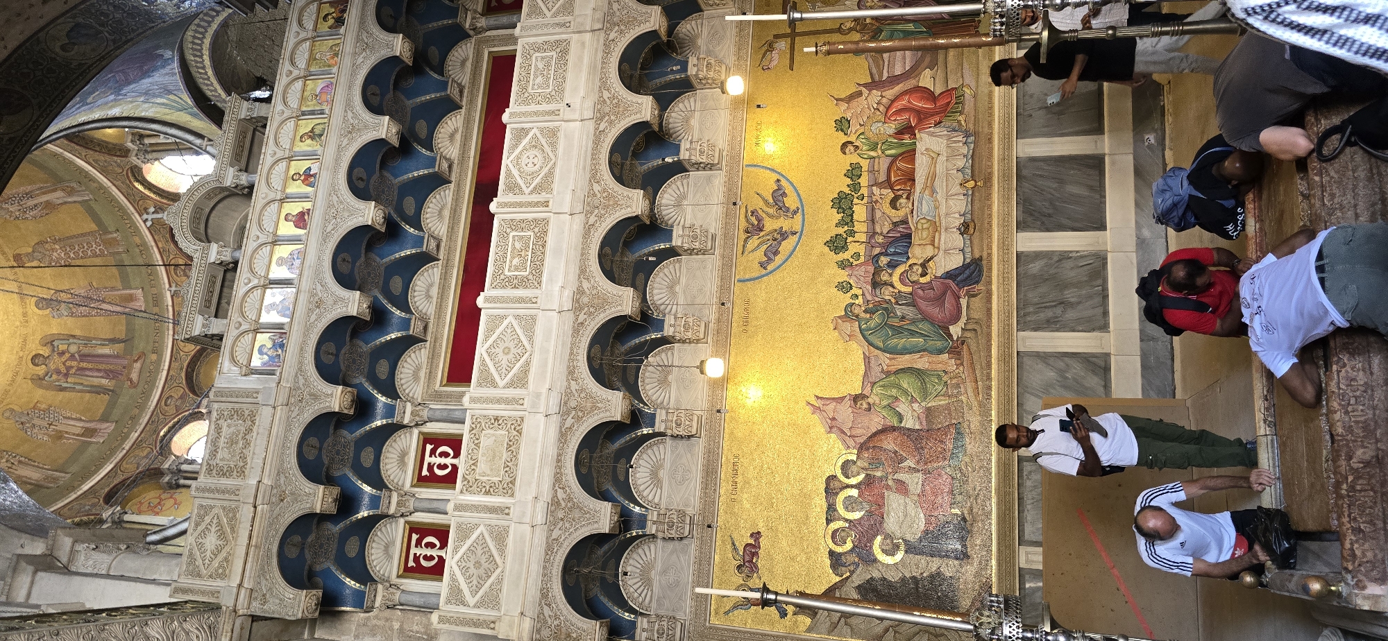 The image captures the interior of the Church of the Holy Sepulchre in Jerusalem, Israel, a significant Christian pilgrimage site. In the foreground, several individuals are seen in prayer, with some kneeling and touching a stone structure, likely the Stone of Anointing. The individuals are dressed in casual attire, including t-shirts and cargo pants, and some have backpacks. A man in a white t-shirt and green cargo pants stands observing while holding a smartphone. Behind them, a large, ornate mosaic depicts a religious scene, possibly the Entombment of Christ, set against a golden background with figures in traditional robes. Above the mosaic, architectural elements of the church are visible, including tiered arches, decorative stonework, and a domed ceiling adorned with religious artwork. The overall scene conveys a sense of devotion and historical reverence within the sacred space.