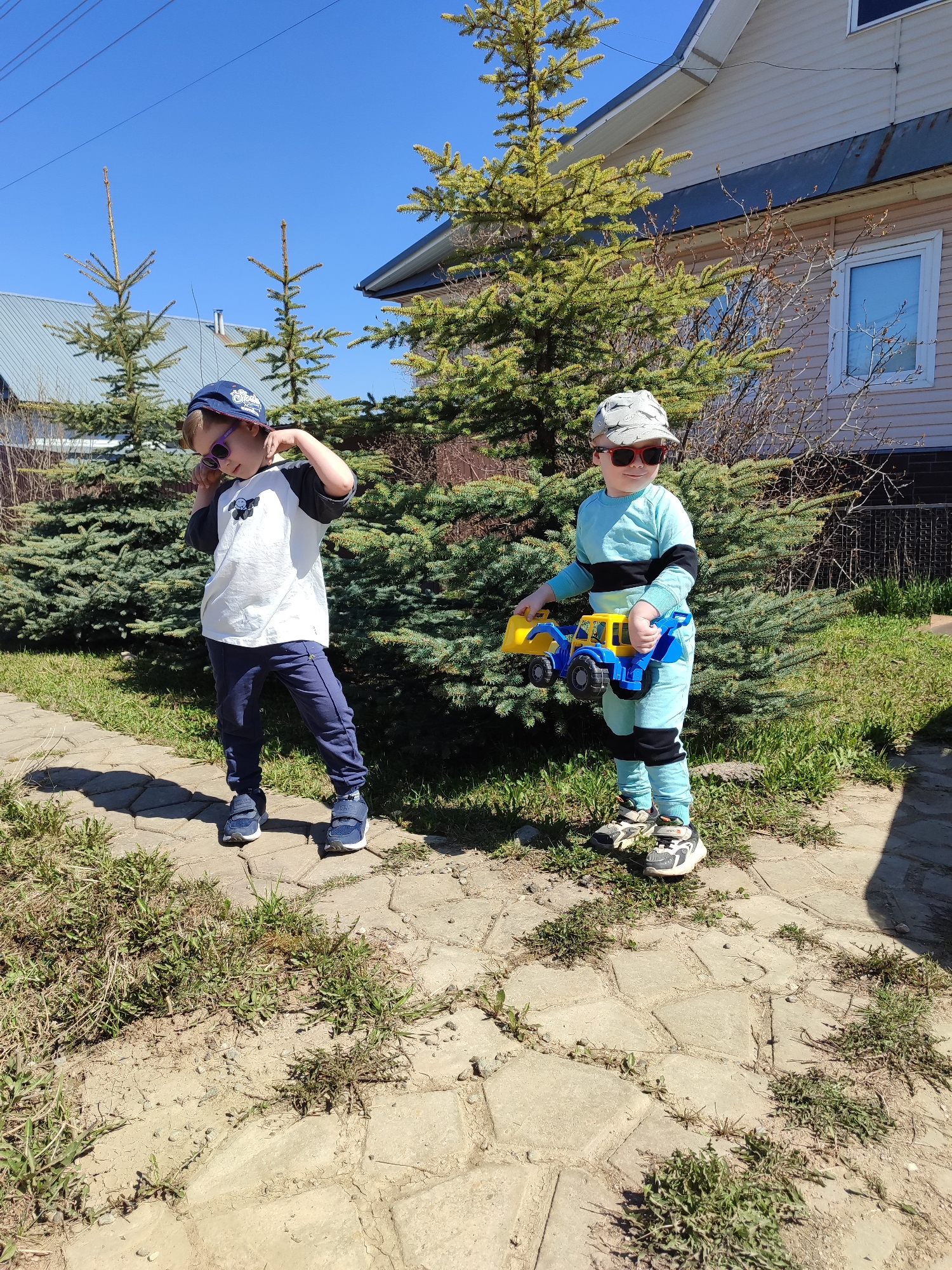 Two young boys stand on a paved walkway in what appears to be a residential area of Malaya Purga, Russia. The boy on the left wears a blue baseball cap, sunglasses, a white t-shirt with dark sleeves, and blue pants, striking a pose with one hand near his head. The boy on the right wears a patterned hat, sunglasses, and a matching light blue long-sleeved shirt and pants; he holds a blue and yellow toy truck. Behind them are several evergreen trees, and in the background is a light-colored, single-story house with a visible window and roof. The sky is clear and blue. The walkway is made of irregularly shaped stones and is bordered by patches of grass and weeds. Electrical wires are visible at the top of the frame.