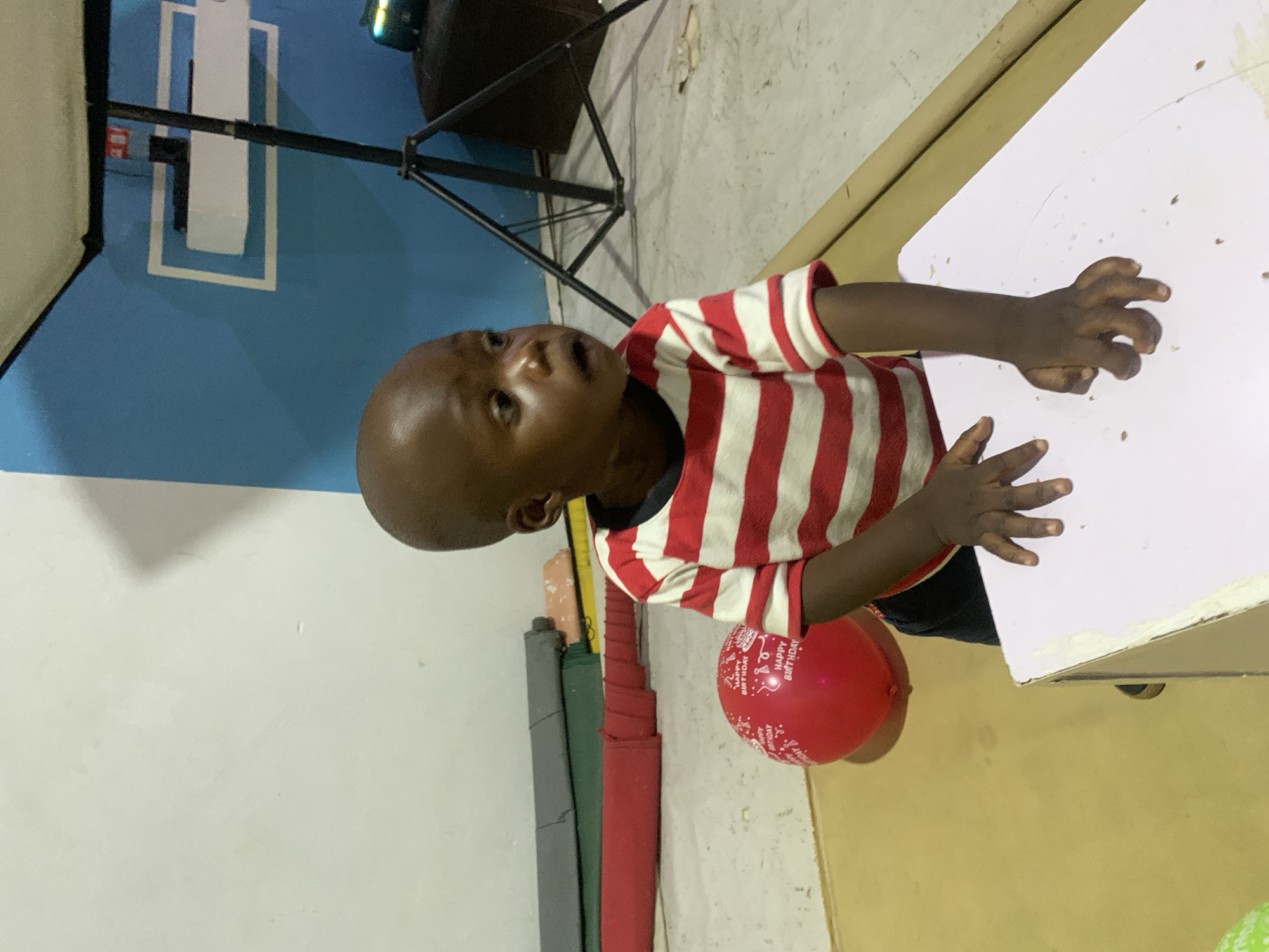 A young child wearing a red and white striped shirt stands next to a white table with what appears to be food scattered on it. The child looks upward, with one hand on the table. A red "Happy Birthday" balloon is near the child. The scene appears to be indoors with a blue wall in the background with what looks like a white painted square with a rectangle inside. This could be in the city of Jos, Nigeria.