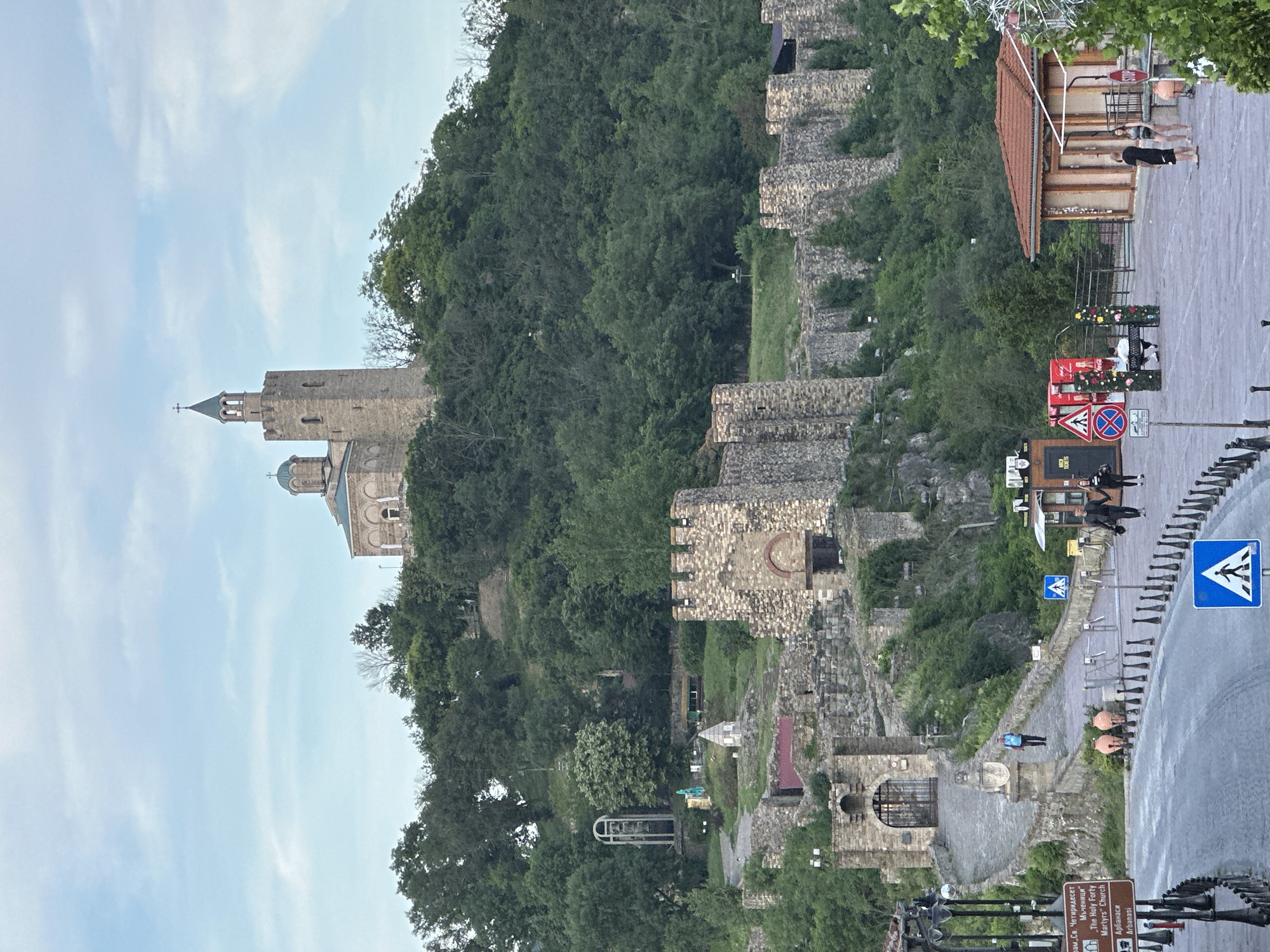 The image shows Tsarevets Fortress in Veliko Tarnovo, Bulgaria. The fortress is situated on a hill and features stone walls, towers, and a church with a dome and a tall spire. The fortress is surrounded by lush green trees. In the foreground, there is a paved area with people walking, a street with traffic signs, and a few buildings, including a small structure with a red tiled roof. The sky is partly cloudy.