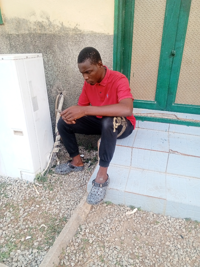 A dark-skinned man sits on tiled stairs in front of a green-framed window. He wears a red polo shirt and black pants with gold embellishments, along with patterned clogs. An air conditioning unit sits to his left on a gravel-covered ground. He looks down, his hands clasped together, appearing contemplative. A concrete curb separates the gravel area from the tiled steps.