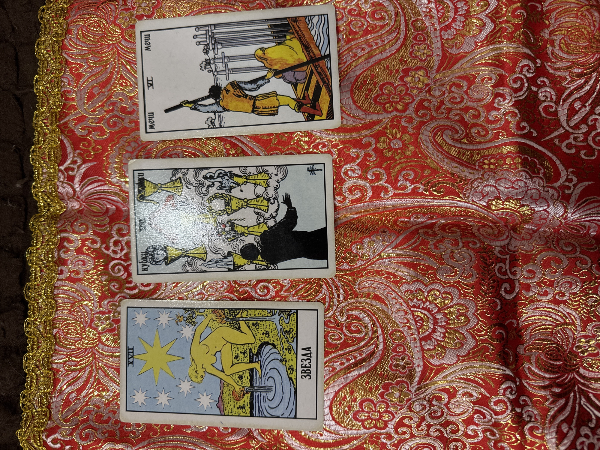 The image shows three tarot cards laid out on a red and gold patterned cloth. The cards are, from top to bottom: Six of Swords, Four of Cups, and The Star. The cards feature writing in a Cyrillic alphabet. The edges of the cloth are lined with a gold trim. The setting is indoors. The location is unknown.