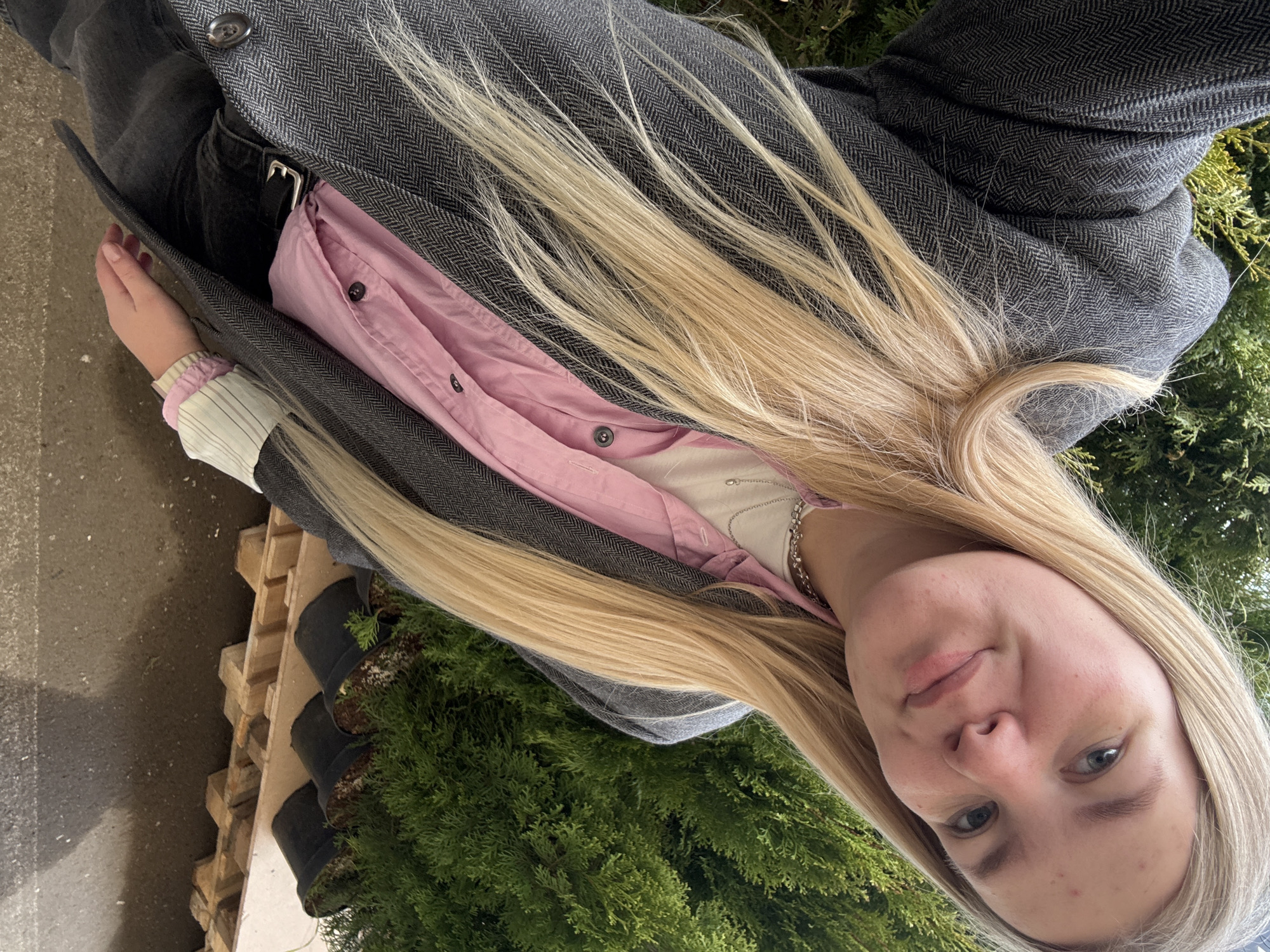 A woman with fair skin and long, blonde hair is facing the camera. She is wearing a grey tweed jacket, a pink shirt, and a white long-sleeved top underneath. She is standing near a stack of wooden pallets with black pots containing small evergreen trees. A section of concrete is visible on the left edge of the frame. The background includes additional evergreen foliage. The location is presumed to be outdoors, possibly at a plant nursery or garden center, but it cannot be conclusively determined to be within the city of Izhevsk, Russia.