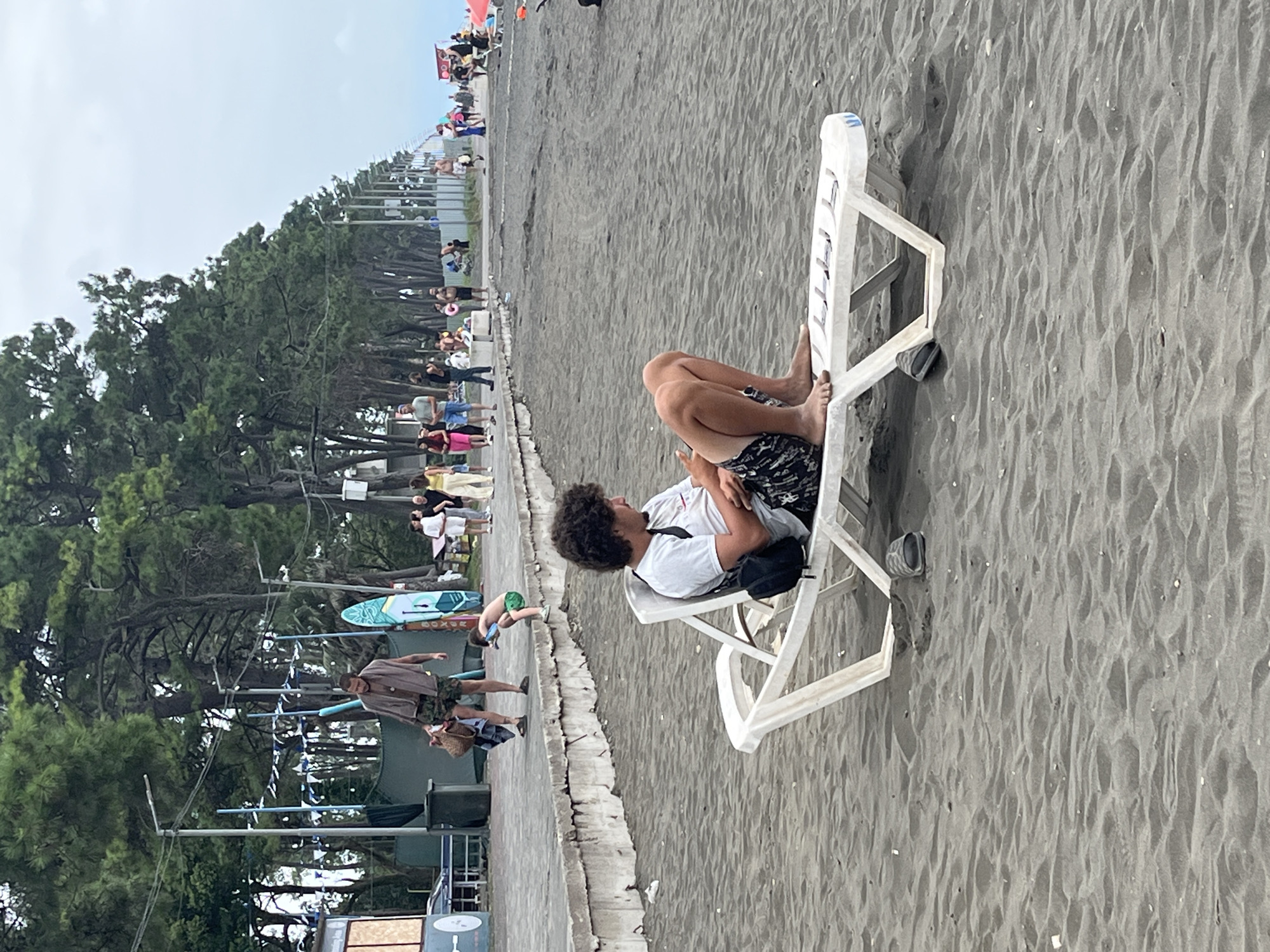 A man with curly dark hair is sitting on a white lounge chair on a dark sand beach in Ureki, Georgia. He is wearing a white t-shirt and patterned shorts and is looking at his phone. His feet are bare. A black backpack is behind him on the chair. A dark-colored flip-flop is on the sand near the chair. In the background, there is a paved promenade with many people walking and standing. There are trees lining the promenade, and further in the distance, more trees and buildings. The sky is overcast.