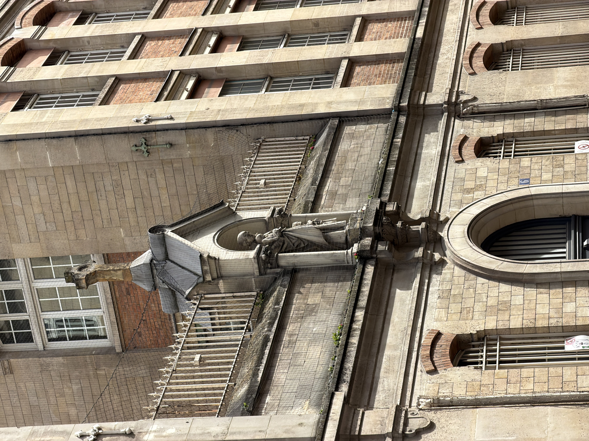 The image shows the exterior of a building in Paris, France, specifically focusing on a section featuring architectural details such as stone carvings, a statue under a small structure with a grey roof, a gargoyle-like projection, and various windows and other openings in the façade. The building appears to be constructed primarily of stone and brick and is covered in some areas by netting. Architectural elements include ledges, cornices, and other decorative features. There is no indication of people in the scene.