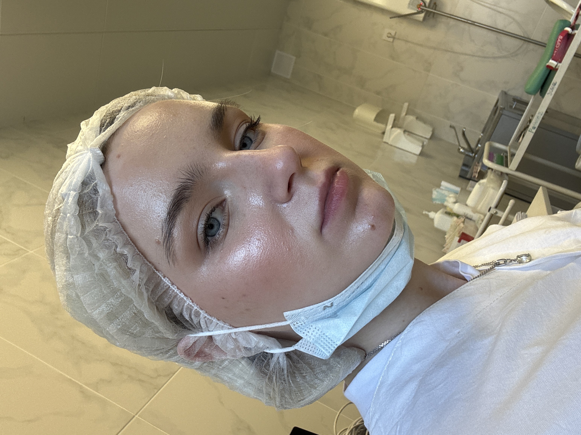 A person wearing a white surgical cap and a blue surgical mask is lying on their back. The person has fair skin, light eyebrows, and blue eyes. They are wearing a white shirt and a silver necklace. The background appears to be a clinical or medical setting, with tiled floors and walls, surgical equipment, and other medical supplies visible in the background.
There is no indication of the image location being in the city of Saint Petersburg, Russia.