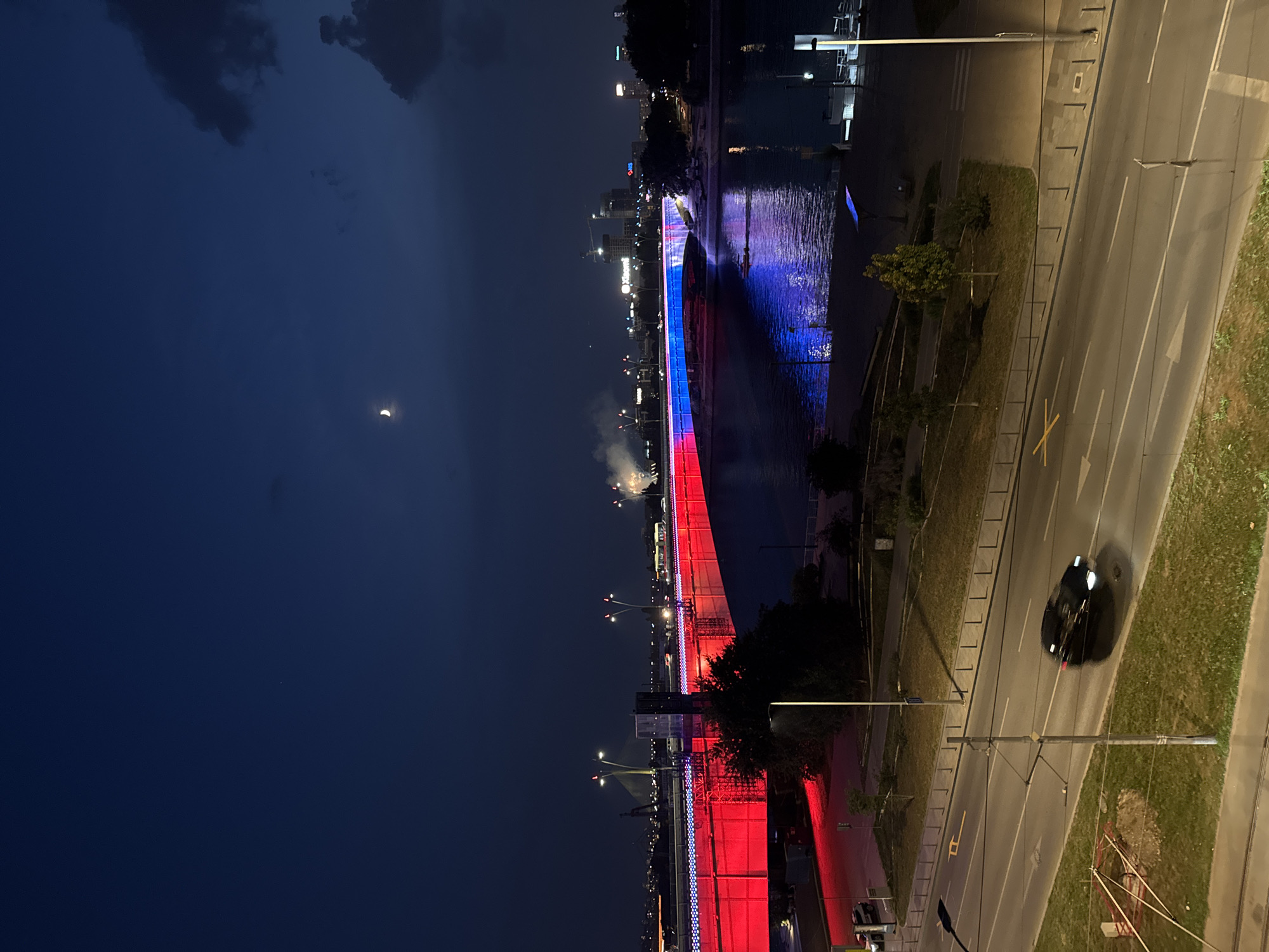 The image shows a night-time view of the Branko's Bridge in Belgrade, Serbia, illuminated with red and blue lights. The bridge spans the Sava River, and its lights are reflected on the water. In the foreground, a dark-colored car is driving on a road. The sky is dark with a crescent moon and some clouds. In the background, city lights and buildings are visible, along with some fireworks or distant lights. The overall scene depicts an urban landscape at night with illuminated infrastructure.