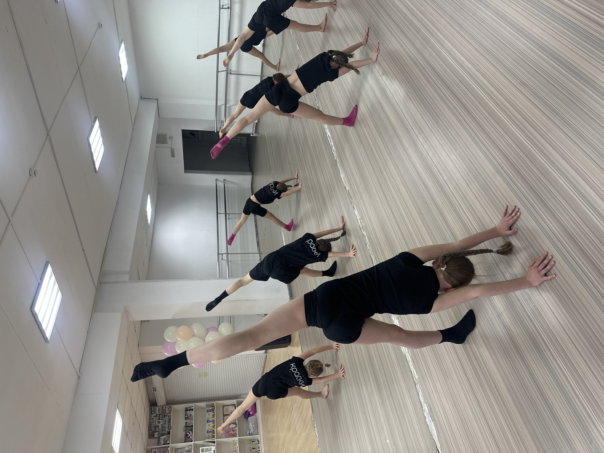 The image shows a dance studio, likely in Votkinsk, Russia, given the writing on the shirts. Seven young dancers, all wearing black shorts and T-shirts with the word "Краски" on the back, are positioned in a line, performing a stretching exercise. They are in a wide stance, with their hands placed on the floor in front of them. Some wear black socks, while others have pink ones. The studio has a light-colored wood-grain floor. A ballet barre is visible in the background. The walls are white, with rectangular ceiling lights. At the back left is a display cabinet. A group of balloons in pastel colors is adjacent to it. A doorway is located further back.