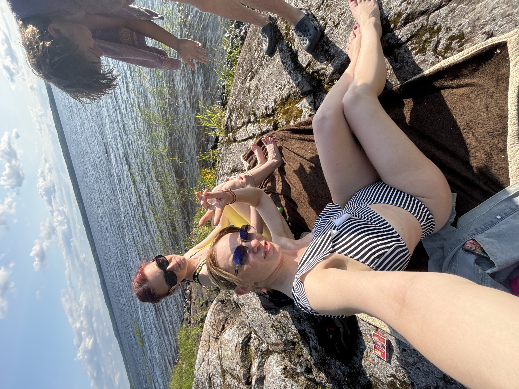 A woman in a black and white striped bikini is taking a selfie on a rocky shore with a lake in the background. She is wearing sunglasses and her legs are crossed. Beside her, another woman in a yellow bikini and sunglasses is sitting and making a peace sign. A third person, partially visible, is standing to the right, their hands reaching out. The scene appears to be a sunny day with a few clouds in the sky. The location is Shuiskaya Chupa, Russia.