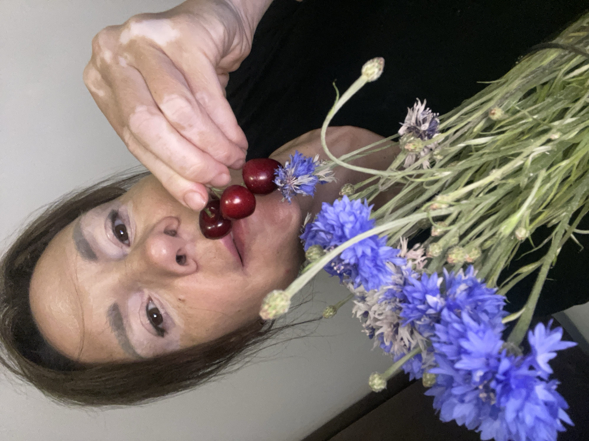 A woman with vitiligo is holding three cherries to her lips, with a bouquet of blue cornflowers in the foreground. She is looking at the camera. The background is a plain, light-colored wall. The image was taken in Sochi, Russia.
