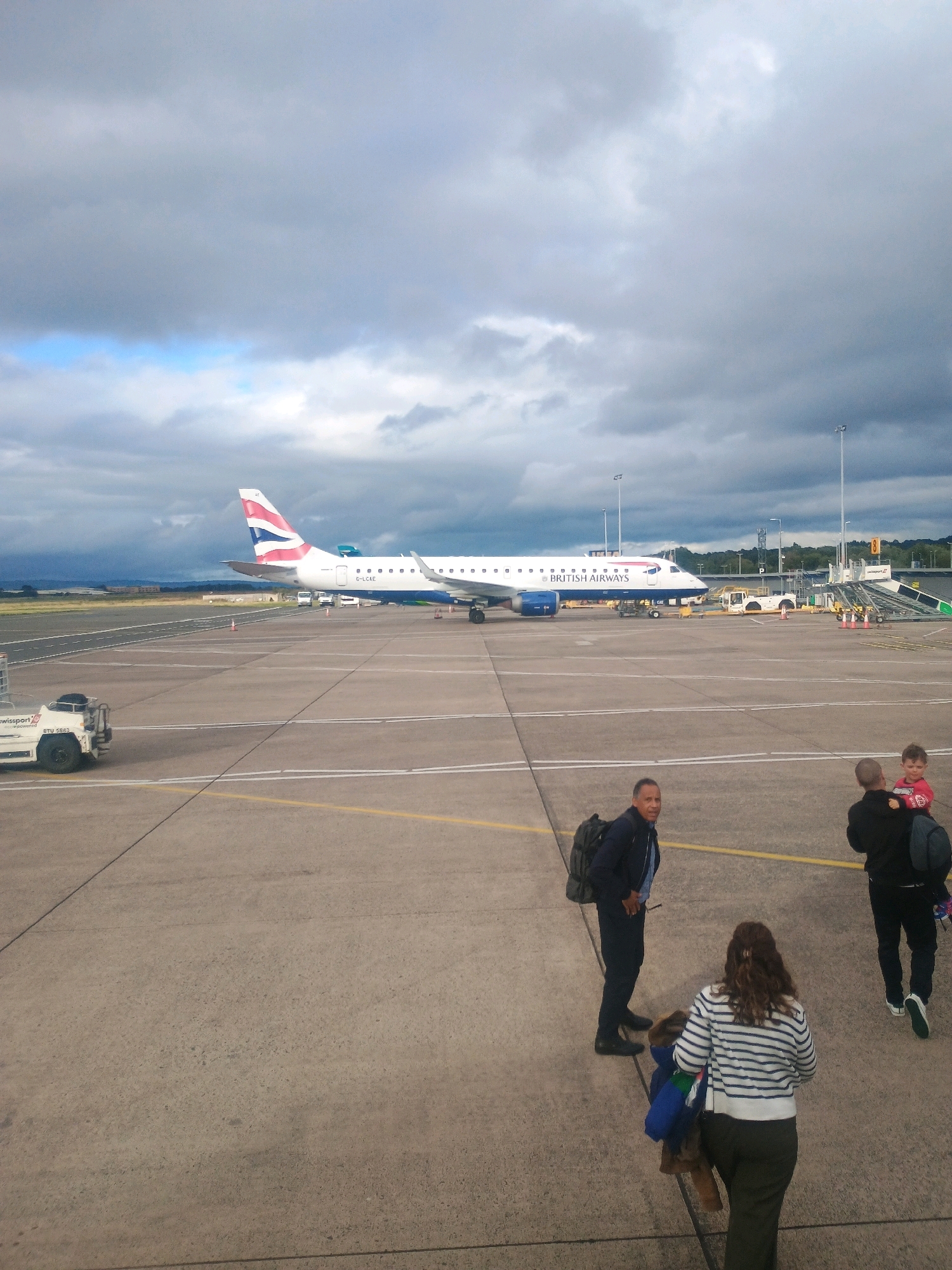 The image depicts an airport tarmac with a British Airways airplane parked in the background. Several people are visible in the foreground. On the left, a man with a backpack is looking towards the camera. In the center right, a woman with long brown hair, wearing a striped shirt and dark pants, is walking away from the camera, carrying a coat. To her right, a man is carrying a child on his shoulders, and another child is walking beside them. The tarmac is a light gray concrete surface with yellow and white lines marking lanes. The sky above is overcast with dark gray clouds. In the far background, trees and buildings can be seen. The scene suggests an active airport environment, possibly during boarding or deplaning.