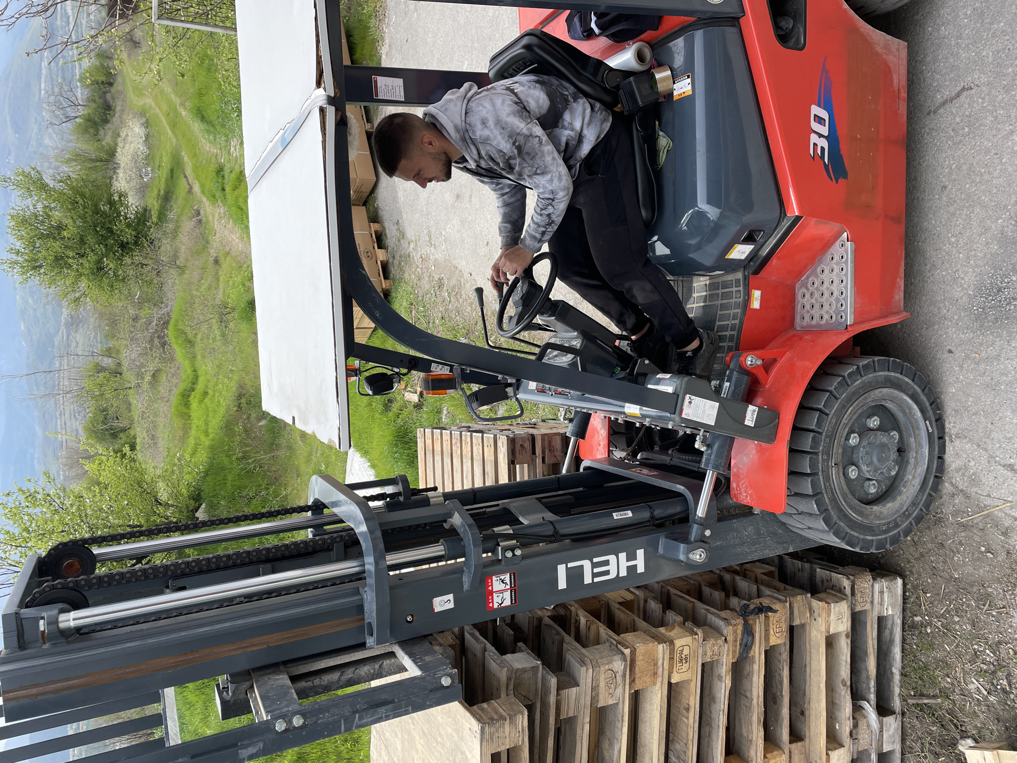 A man is operating a red HELI forklift in an outdoor environment. The forklift is positioned on a stack of wooden pallets. The man is seated in the driver's seat and appears to be manipulating the controls. In the background, there is a building, and beyond that, a landscape with hills and greenery. The location is Asenovgrad, Bulgaria.