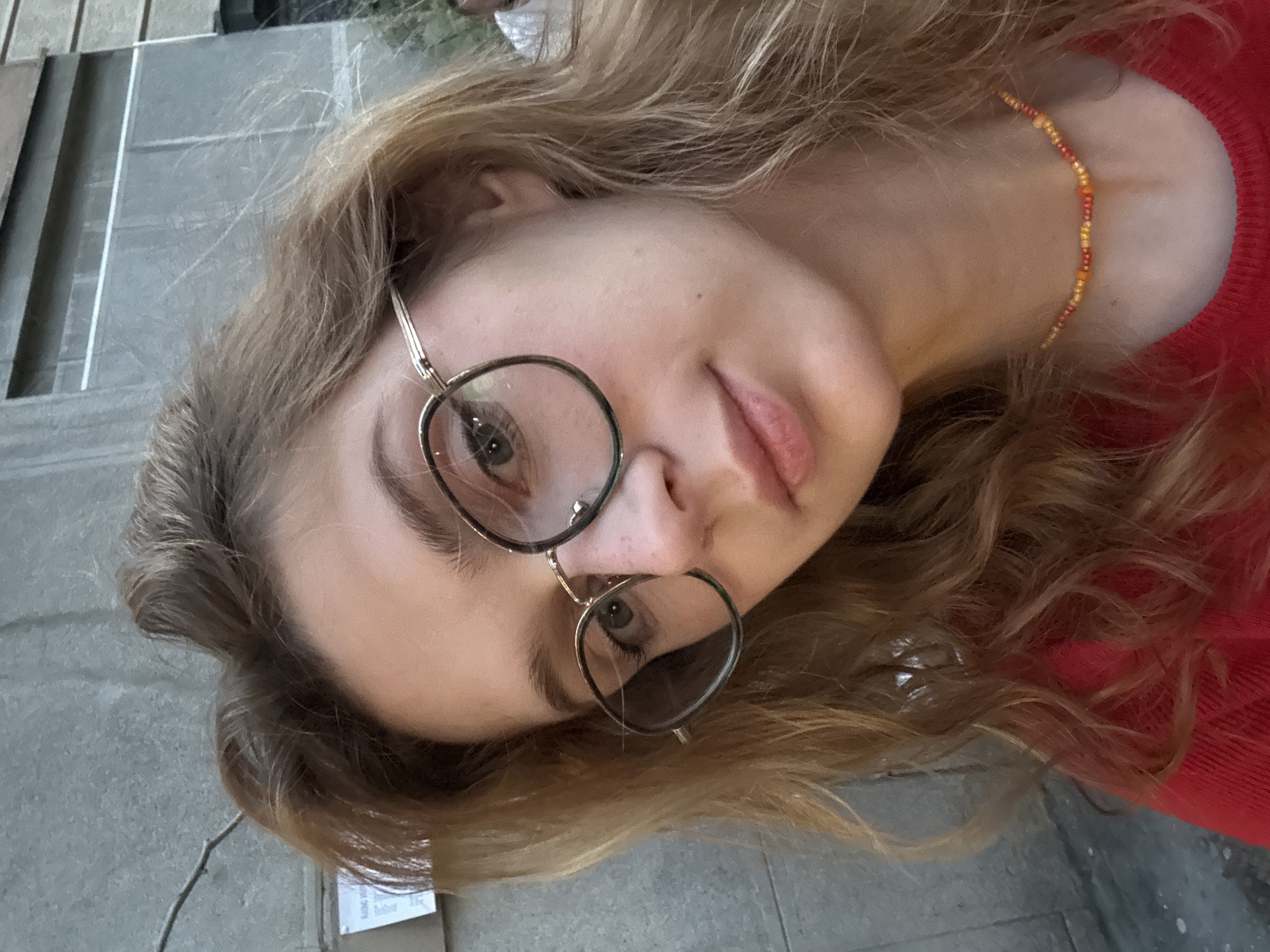 A young woman with light brown, wavy hair, wearing round eyeglasses, is the primary subject of the close-up portrait. She is tilted to the side, her face angled towards the camera. She has fair skin, light eyebrows and visible light eyelashes. She is wearing a red top and a multi-colored beaded necklace. The background includes gray concrete surfaces, suggesting an outdoor urban setting. The exact location in Belgrade, Serbia, cannot be determined from the visual information provided.