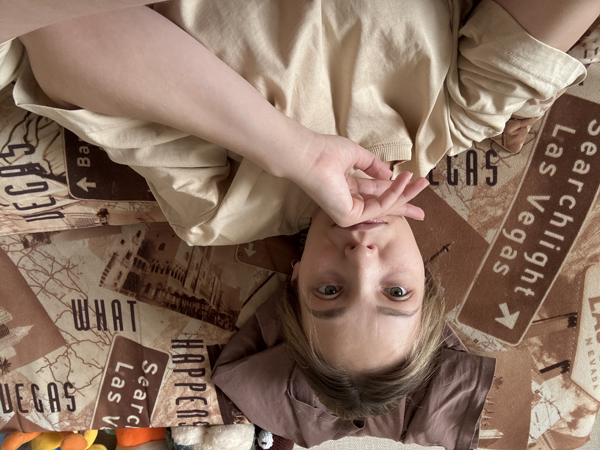 The scene is set indoors within the city of Chaikovsky, Russia. The image captures an overhead selfie of a young adult female with light brown hair and green eyes, possessing a fair complexion. She is attired in a light beige or tan short-sleeved t-shirt with a ribbed crew neck. Her right hand is positioned with fingers gently touching her chin and lower lip area. She maintains a neutral expression while looking directly at the camera. She is lying on a surface covered by a patterned textile, likely a blanket or duvet, featuring a brown and tan color scheme. The pattern incorporates various Las Vegas-themed elements, including road signs with text such as 'Searchlight Las Vegas' and 'WHAT HAPPENS,' alongside images of buildings and a stylized map. Behind her head, a crumpled brown fabric garment is visible. To the upper right, partially obscured, are several plush toys or decorative items of varying colors, including white, orange, yellow, and blue/green. The immediate environment is an uncluttered indoor space.