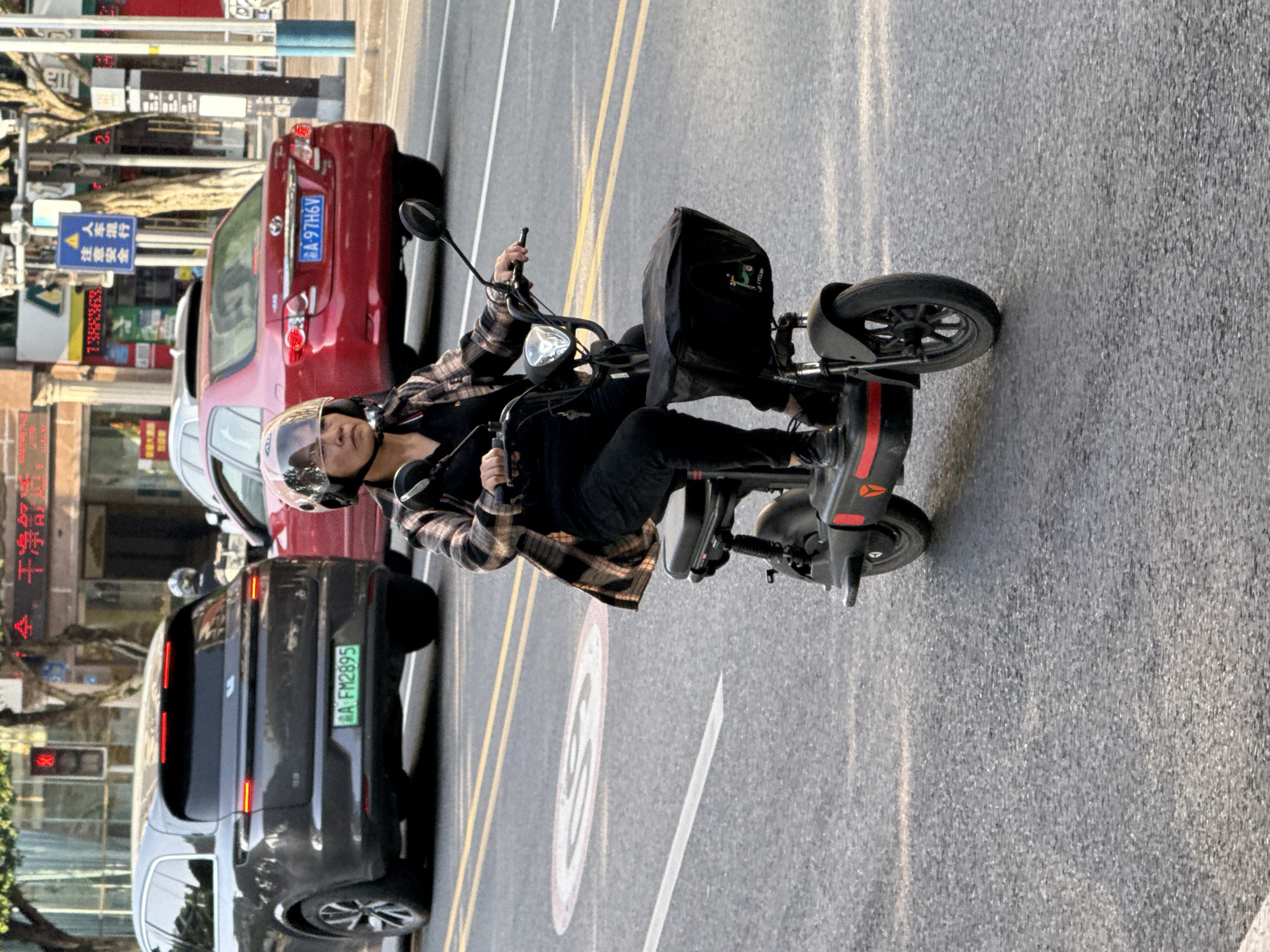 A person wearing a helmet with a clear visor and a plaid shirt rides a black electric scooter on a paved street in Jiangjin, China. The scooter has red accents and a black basket or bag mounted at the front, which features the word "CYCLEY" and a small graphic. The rider's expression is neutral as they navigate the road.
Behind the scooter, a red sedan with license plate "A-97H6V" is visible, followed by a dark grey SUV with a green license plate "A FM2895". The street features yellow double lines separating lanes and a circular speed limit sign painted on the road showing "50". In the background, buildings with storefronts, trees, and street signs are visible. A blue sign in the distance displays Chinese characters, which translate to "人车混行" (people and vehicles mixed) and "注意安全" (pay attention to safety). Another electronic display partially shows "全,干净舒适," and a traffic light countdown "2". The scene appears to be taking place on a clear day, with bright sunlight casting shadows.