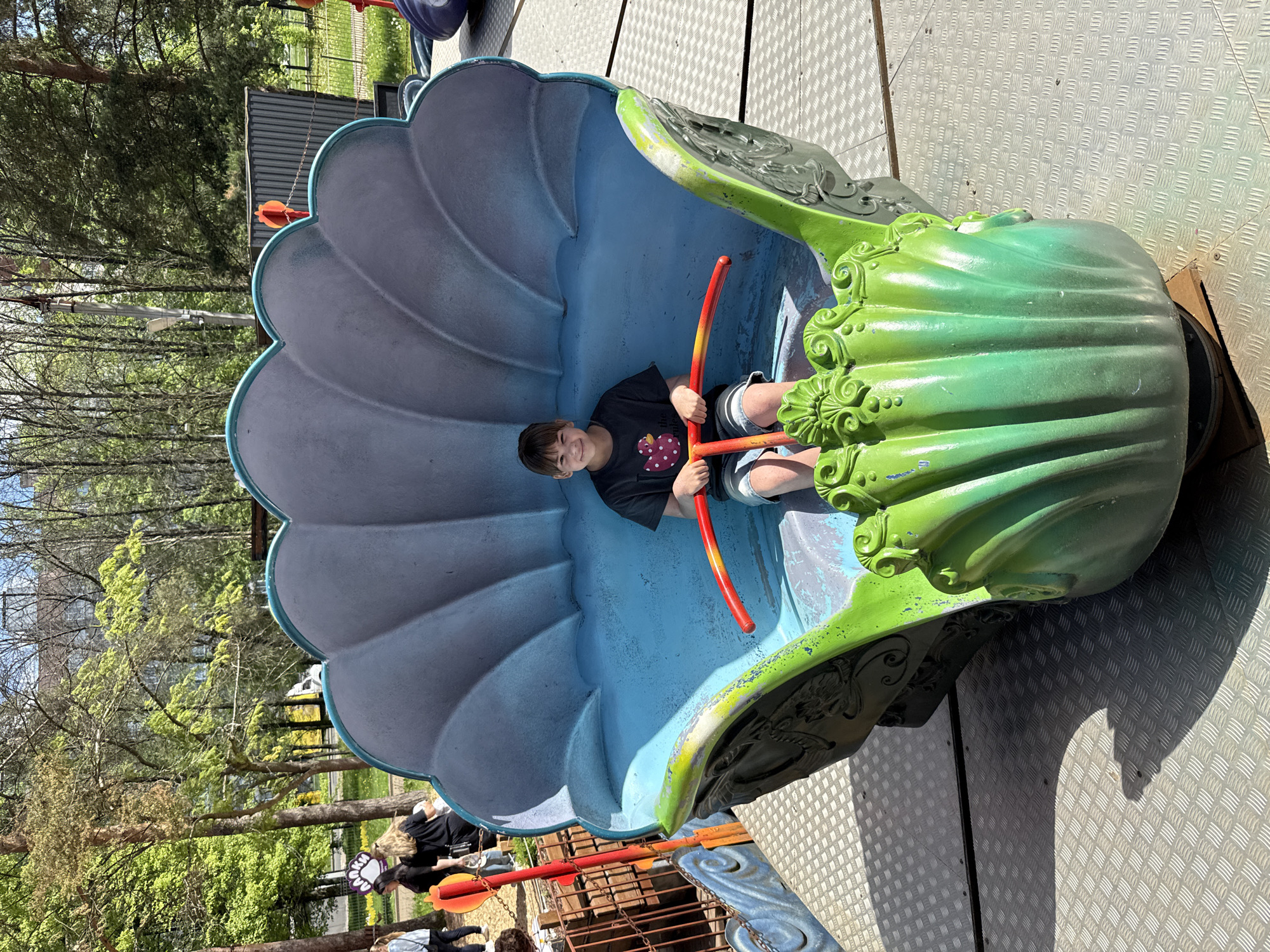 A fair-skinned child sits within a large, clam-shaped ride at what appears to be a playground. The child, wearing a dark shirt with a graphic and shorts, holds two orange-red bars, presumably part of the ride's mechanism. The interior of the clam is predominantly blue, while the outer shell is a gradient of green fading into grey. The ride sits on a patterned metal surface. Outside the immediate structure, one can see trees and other playground equipment, suggesting an outdoor recreational space. The presence of other people in the background near the base of the structure indicates that the area is being used by the public. It is assumed to be located at Rusanovo, Russia.