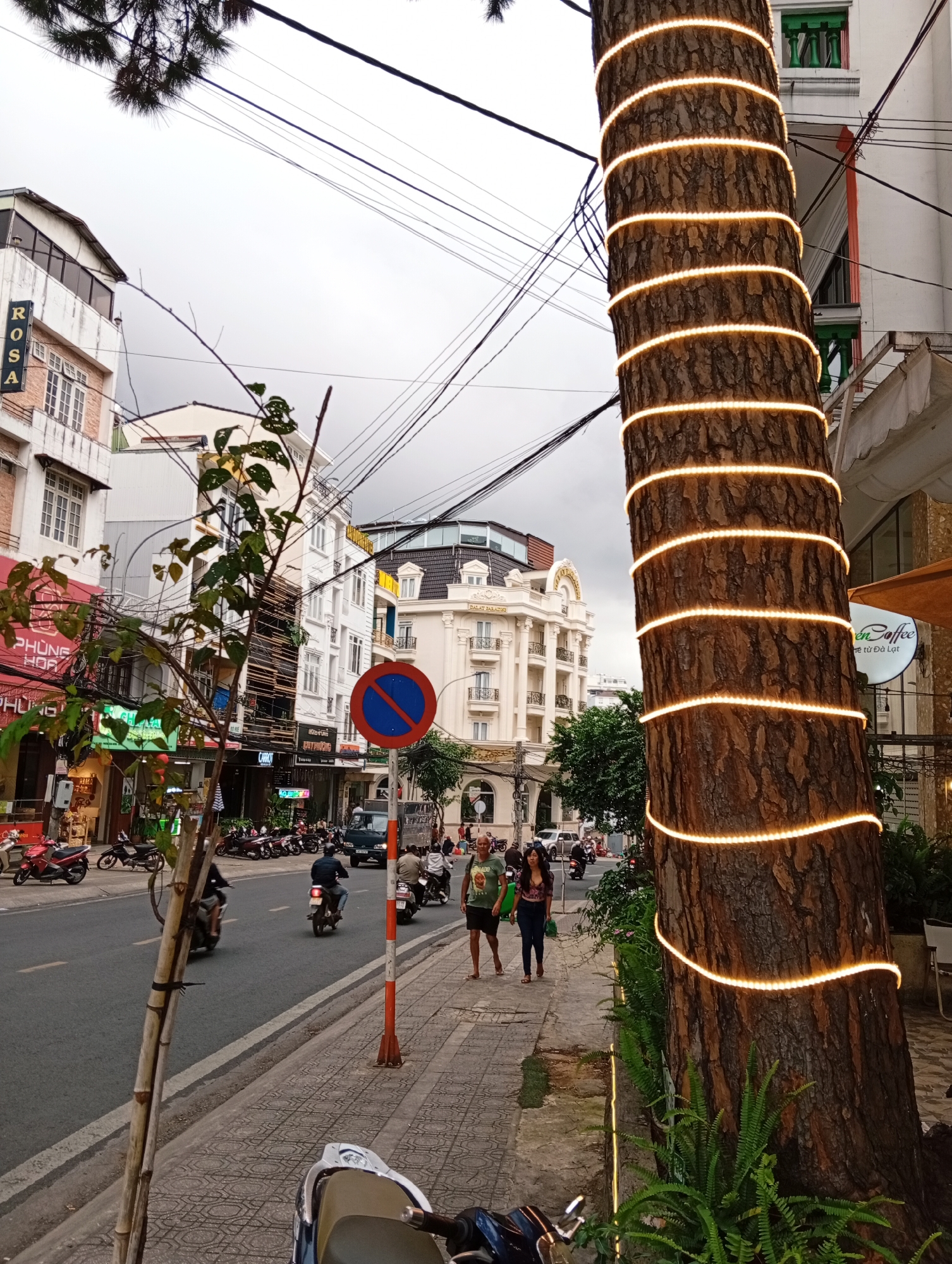 A tree trunk wrapped in a spiral of warm-toned LED lights is the primary focus of the image, situated on the right side. The tree is located on a sidewalk adjacent to a street in Da Lat, Vietnam. The street is busy with traffic, including motorcycles, a truck, and cars. Pedestrians walk along the sidewalk, and various shops and buildings line the street. The architecture of the buildings suggests a commercial district. The sky is overcast, and the overall lighting indicates it's daytime, possibly late afternoon or early evening, given the artificial lights on the tree. A sign with a blue circle and a red diagonal line, indicating a traffic restriction, is visible on the sidewalk. The scene captures a typical urban environment in Da Lat.