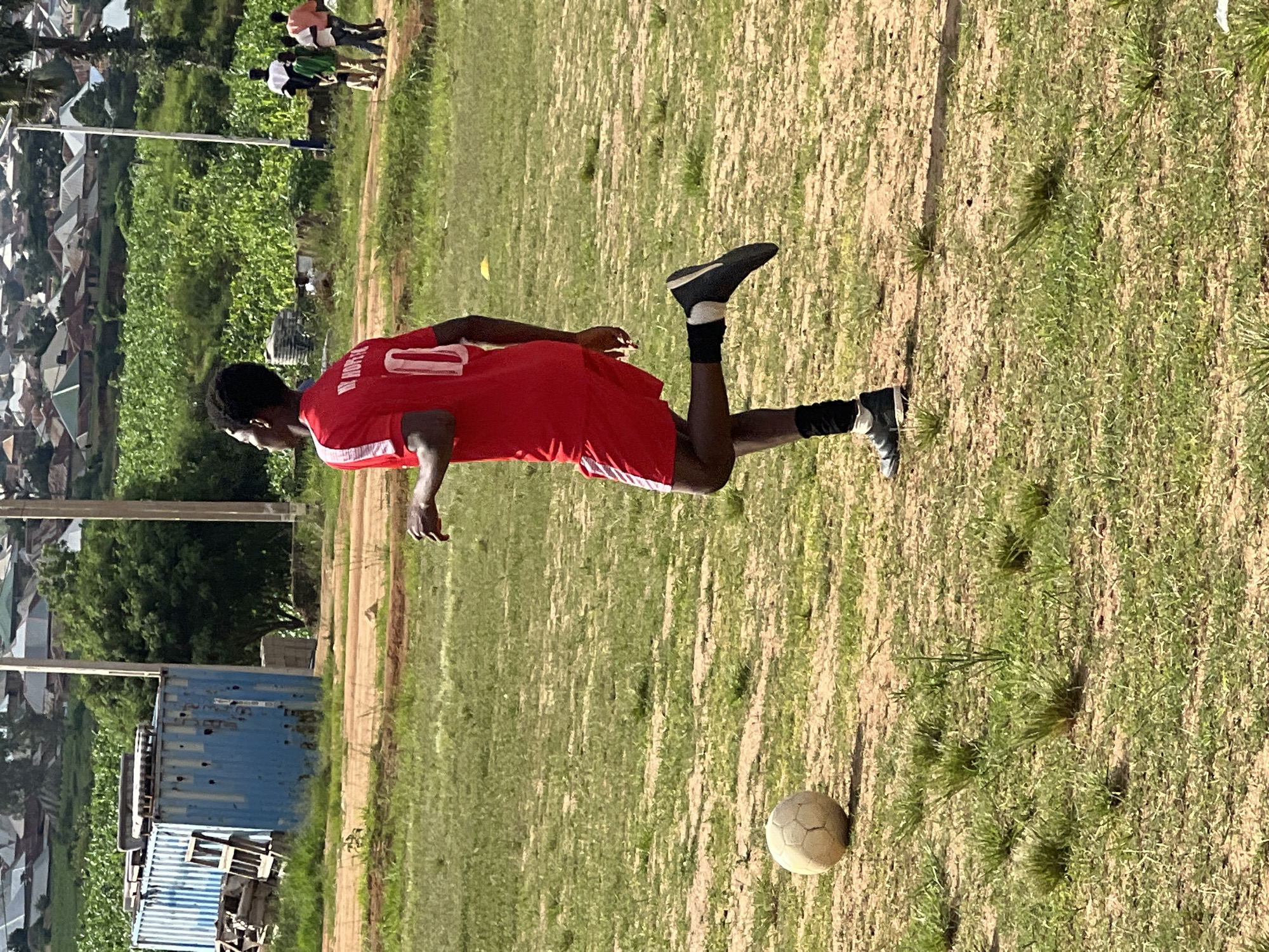 An outdoor, medium-shot, eye-level, horizontal photograph depicts a young Black male soccer player in a red jersey and red shorts with white stripes on the sides, and black soccer cleats with white socks on a dirt and grass field. He is in mid-stride, with his right leg extended backward and his left leg bent at the knee. He appears to be running or dribbling. The jersey has "M. HOPE" and the number "10" printed in white on the back. A tan soccer ball is visible on the grass to the left of the player. In the background, there is a blue shipping container, some wooden debris, a patch of green crops, and a densely populated residential area with buildings featuring corrugated metal and tiled roofs, characteristic of the city of Jos, Nigeria. Several other people, appearing to be mostly young men and boys, are visible in the background, some of whom are also wearing athletic clothing. The sky is bright and appears to be clear. The overall scene suggests a casual soccer game or practice session taking place in a semi-urban environment.