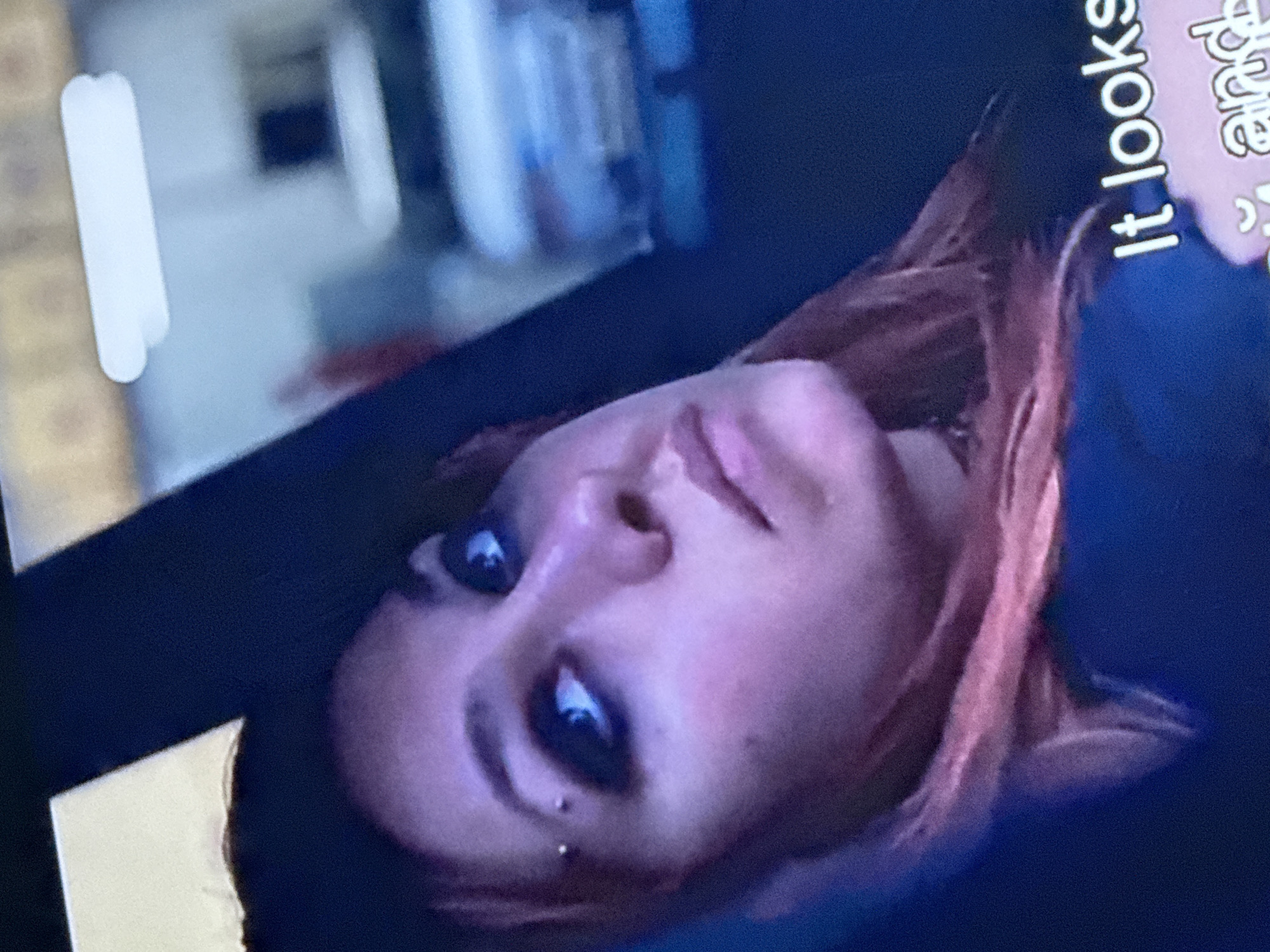 The image shows a person with pinkish skin and dark eyes, possibly female, with a piercing on the left side of their lower lip. The person has reddish-brown hair. A portion of text reading "It looks and" is visible to the right of the person's head. The background appears to be an interior space, possibly a room with a shelf-like structure. There is not enough information to identify the location as Mytishchi, Russia.