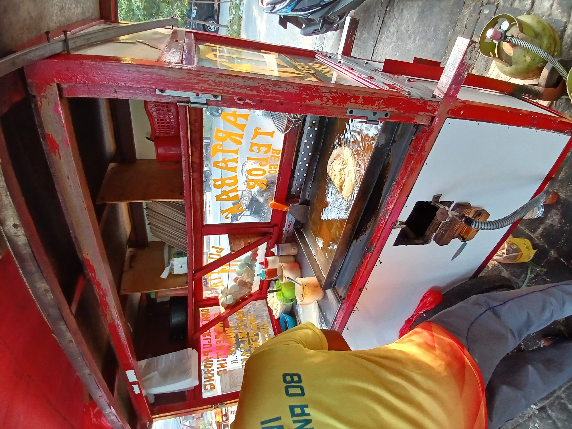 In a sunlit outdoor scene in Bekasi, Indonesia, a food vendor, identified by the text "INA 08" on their bright yellow shirt, is actively preparing food. They stand behind a red and white food cart, which is equipped with a cooking station featuring a griddle submerged in hot oil. A partially cooked food item, likely an egg-based dish, is visible in the bubbling oil. The cart's overhead shelf displays various ingredients and cooking utensils, including bowls of shredded vegetables, seasoning bottles, and a small whisk. Text on the cart's glass panel, written in yellow capital letters, advertises "MARTABAK TELOR" and "TELOR PADANG," indicating the type of food being sold. A propane tank is positioned near the base of the cart, connected to the cooking apparatus via a hose. The surrounding environment suggests a street-side location, with pavement and a glimpse of parked vehicles and vegetation in the background.