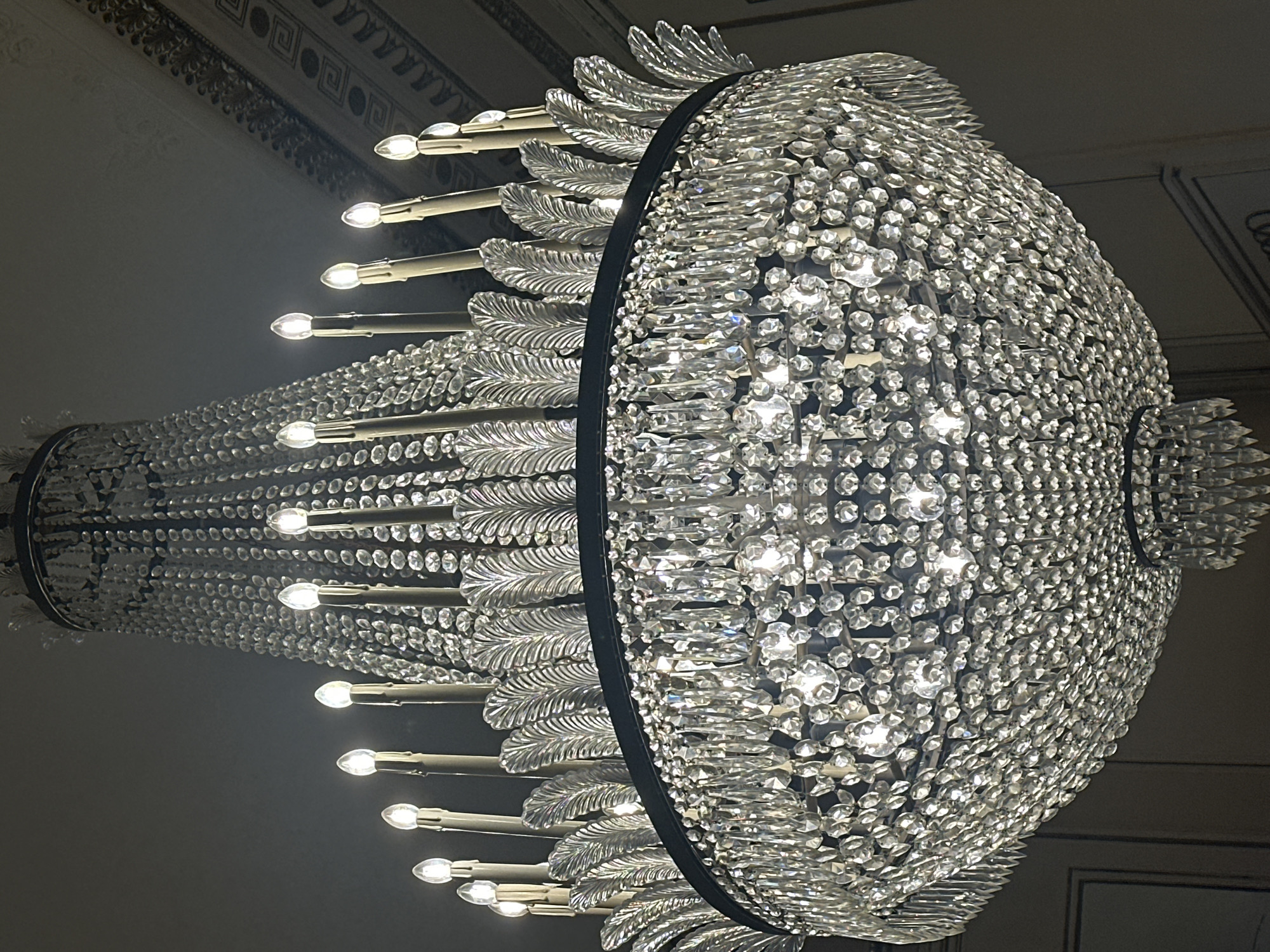 A large, ornate crystal chandelier hangs from a ceiling in a dimly lit interior. The chandelier features multiple tiers of faceted crystal prisms and teardrop-shaped pendants that catch and reflect the light from numerous candle-shaped bulbs. Decorative glass or crystal leaves are interspersed among the upper tiers. The ceiling above the chandelier has intricate moldings and decorative detailing. The background walls are a muted color, possibly beige or gray, with faint vertical lines suggesting paneling or doors. The image is taken from a low angle, looking upwards towards the chandelier. The location is identified as Unknown, Unknown.