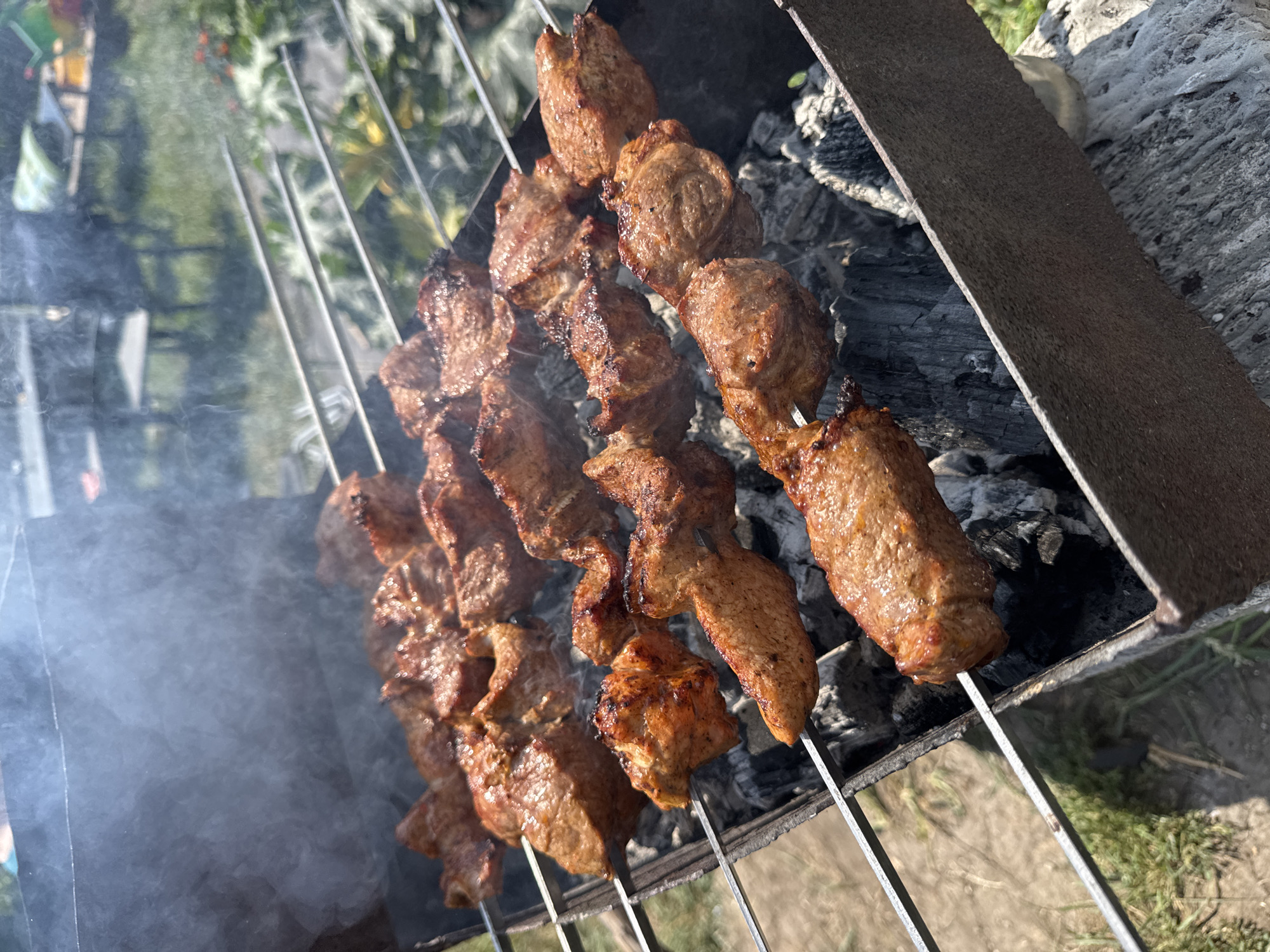 In the village of Uzyukovo, Russia, on a sunny day, several skewers of meat are being grilled over hot coals in a portable barbecue. The meat, cut into bite-sized pieces, is browned and slightly charred from the heat. Smoke rises from the grill, indicating the cooking process is in progress. The barbecue is placed outdoors, with a grassy area and some greenery visible in the background. The scene suggests a casual outdoor meal or gathering.