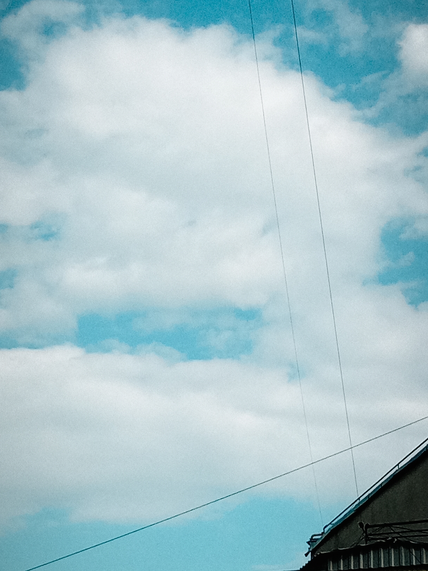 The image shows a sky with clouds and electrical wires against a backdrop of buildings. The clouds are white and fluffy, and the sky is a bright blue. The electrical wires are thin and dark, stretching across the frame from top to bottom. The buildings are dark and appear to be made of concrete. The scene is likely a typical urban landscape in Chișinău, Moldova, where such overhead wires are common.