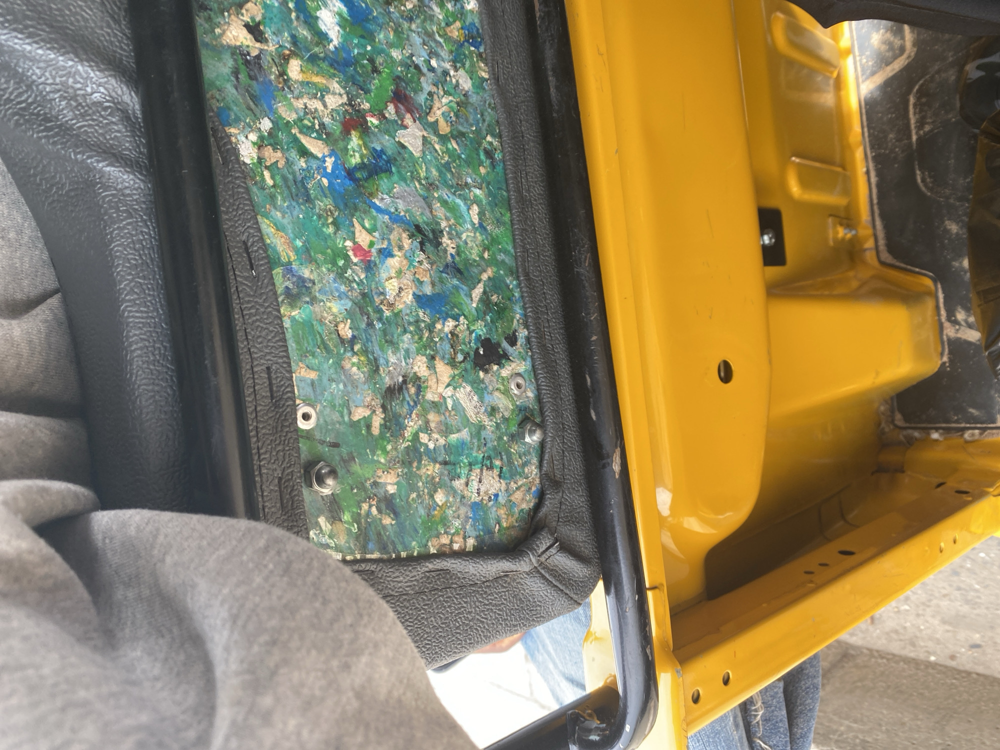 This media file is a close-up, interior shot of a yellow vehicle, likely a utility vehicle or truck, in Jalingo, Nigeria. The focus is on the driver's side door panel and the surrounding interior. The door panel itself is partially removed, revealing a multi-colored material underneath, composed of what appears to be shredded or recycled material in shades of green, blue, red, and tan. This material is held in place by a black textured plastic trim.
To the left of the door panel, a grey, textured fabric, possibly part of a seat or a garment, is visible. A black metal bar, part of the vehicle's frame or a handle, runs vertically along the right edge of the door panel. The right side of the image shows the bright yellow painted interior of the vehicle's body, with some visible screws and mounting points. A small black rectangular object with a screw is attached to the yellow surface.
The scene appears to be in an outdoor setting, with ambient light suggesting it is daytime. No specific weather conditions are evident. The overall impression is one of an interior viewed from a low angle, possibly by someone sitting in the vehicle. The condition of the door panel suggests it might be undergoing repair or has been modified. There is no visible text in the image.