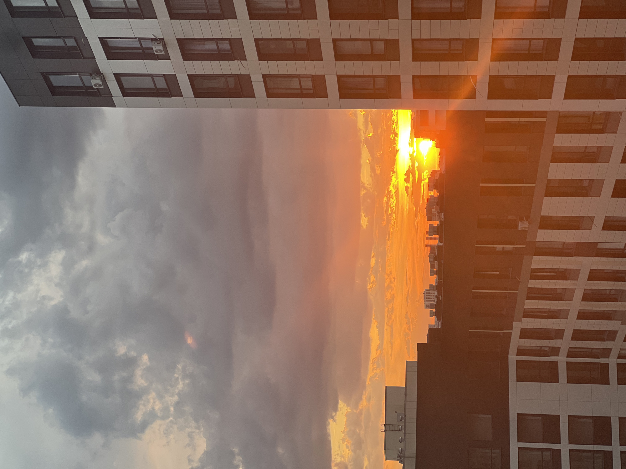 A sunlit sunset over a cityscape in Astana, Kazakhstan. The image captures a sky ablaze with orange and yellow hues, with scattered dark clouds creating a dramatic contrast. In the foreground, modern high-rise buildings with repeating window patterns dominate the right and lower sections of the frame. The buildings feature a mix of dark and light facades, with some windows reflecting the sunset's glow. The left side of the image shows the tops of other buildings, partially obscured by the setting sun. The overall scene depicts an urban environment bathed in the warm light of a sunset.