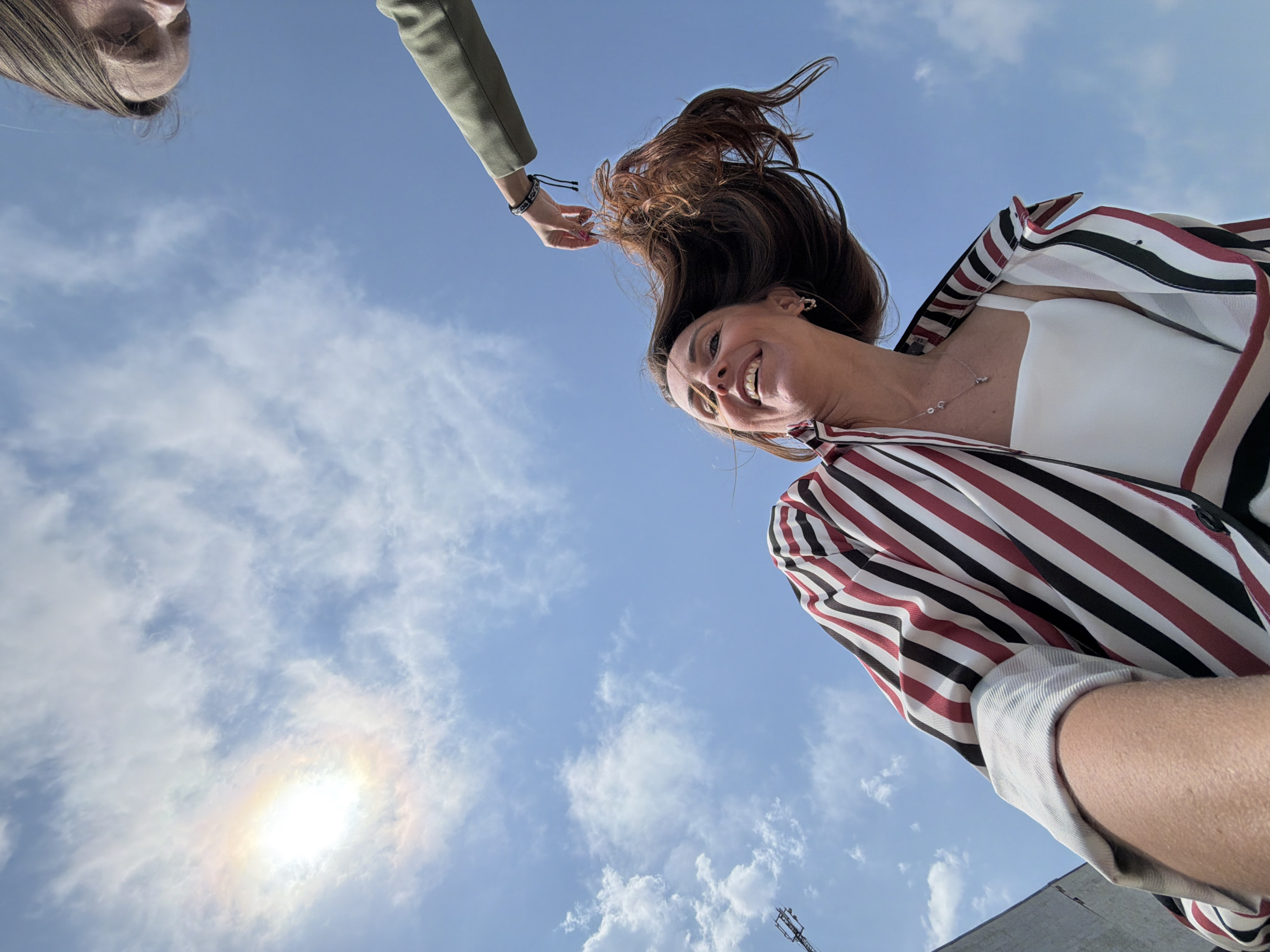 The image shows a low-angle view of two women against a blue sky with scattered clouds and the sun. One woman, seen from the chest up, is smiling with her head tilted back, her hair flying. She wears a white top under a striped shirt of white, black, and maroon stripes. A second arm and hand, belonging to a person not fully visible in the frame, are reaching toward the woman's hair. A third person's face is partially visible in the upper left corner. In the background, parts of a building and a utility pole are visible. The specific location within the city of Birobidzhan, Russia, cannot be determined based on the information provided in the image.