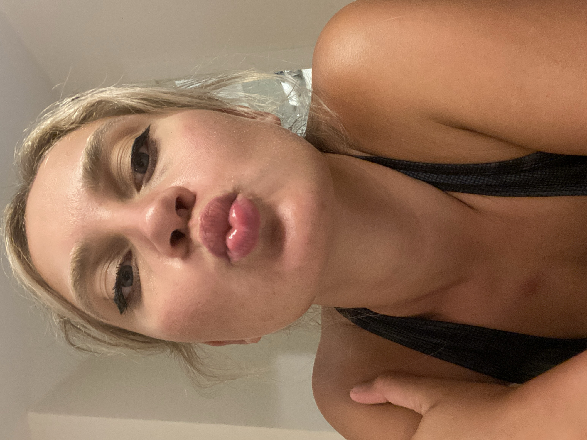 A close-up, low-angle selfie shows a young woman with blonde hair pulled back, puckering her lips towards the camera. She is wearing a dark-colored athletic top. Her eyes are lined with black eyeliner, and her lips are glossy. The background is a plain, light-colored wall. The image appears to have been taken indoors. There is no indication of the location in Aksu, Turkey within the provided image.