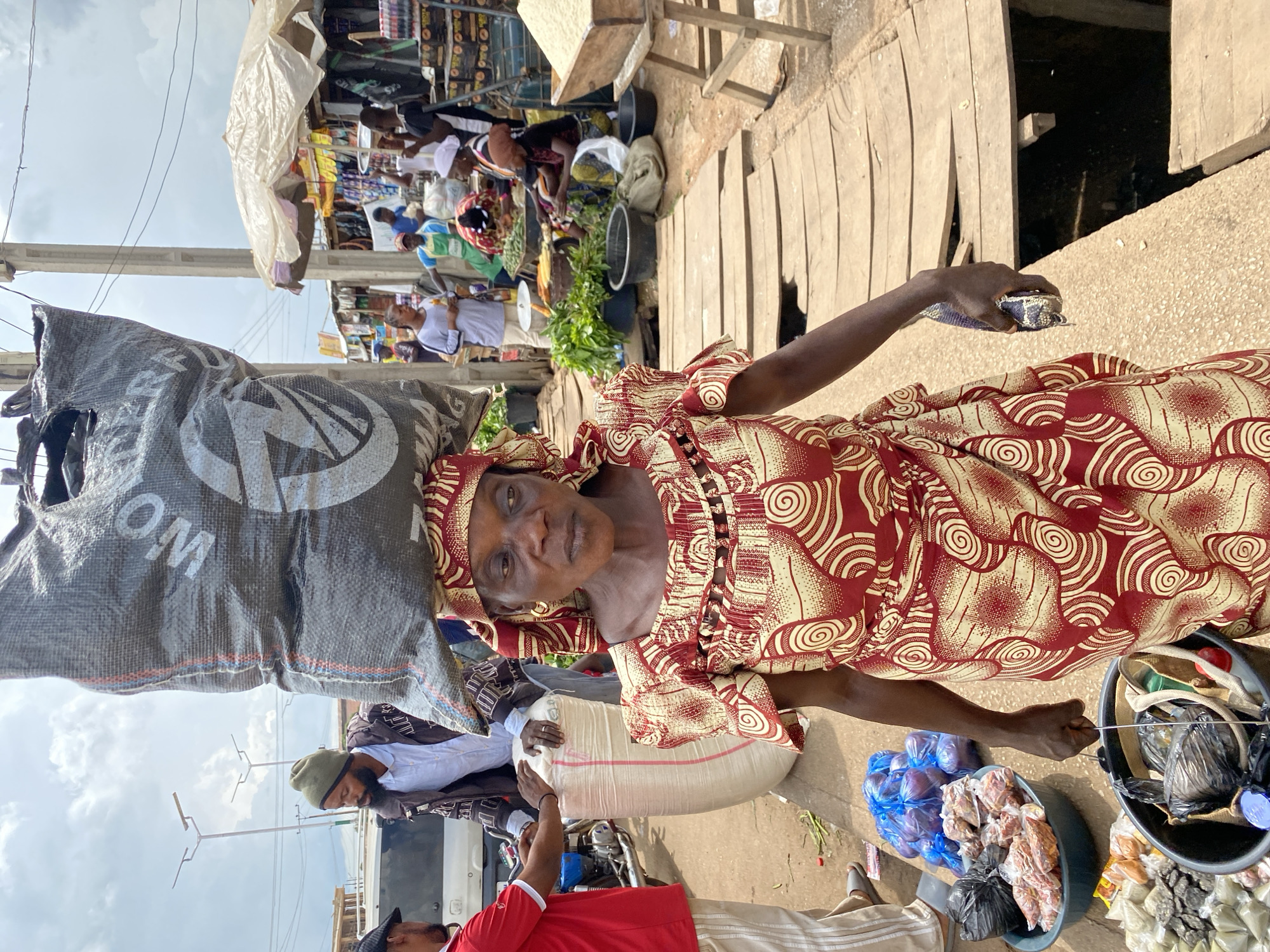 In a bustling outdoor market in Bukuru, Nigeria, a woman with dark skin and wearing a red and yellow patterned dress and headscarf carries a large, dark grey sack on her head. The sack has white lettering, partially visible, reading "MONTERRA". She is walking on a wooden walkway that spans over a dark, open space.
In the background, other people are engaged in market activities. To the left, a man in a red shirt and a beanie is interacting with another man in a brown jacket and white shirt, near a large white sack. Further behind them, a white vehicle is parked.
The market appears to be organized with stalls selling various goods, including produce. Several tarps and umbrellas provide shade over some of the stalls. A wooden stand displays a large mound of rice. In the foreground, to the woman's left, are several basins containing produce, including what appear to be purple onions in blue plastic bags, and other food items in plastic bags and bowls. She is holding a dark object, possibly a phone or wallet, in her right hand. The ground is a mix of dirt and gravel, with the wooden walkway dominating the center of the scene. The sky is overcast with clouds.