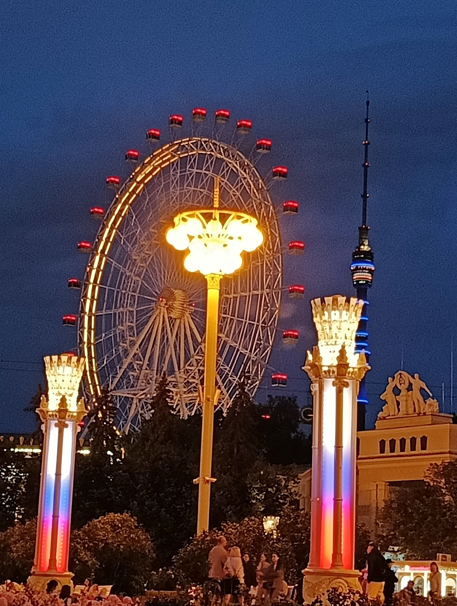 The image shows a Ferris wheel illuminated with yellow lights and red accents on its cabins, set against a dark blue twilight sky. To the right of the Ferris wheel, a tall, dark tower with blue and white lights is visible. In the foreground, two tall, ornate lampposts are lit with vertical bands of red, white, and blue light, reminiscent of the Russian flag. Several people are seen in the lower portion of the image, some standing and some seated, suggesting a public gathering or event. Trees are interspersed throughout the scene. The architectural style of the building with statues on top, as well as the presence of the Ostankino Tower in the background (indicated by its distinctive shape), strongly suggests the location is VDNKh (Exhibition of Achievements of National Economy) in Moscow, Russia.