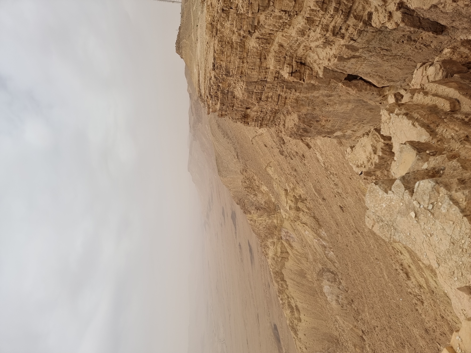 The image shows a rocky desert landscape in Mitzpe Ramon, Israel, possibly a cliff or canyon. The sky is overcast with a thick, hazy cloud cover. The rock formations are primarily light brown and beige, with visible layers and textures indicating sedimentary rock. The landscape is arid and barren.