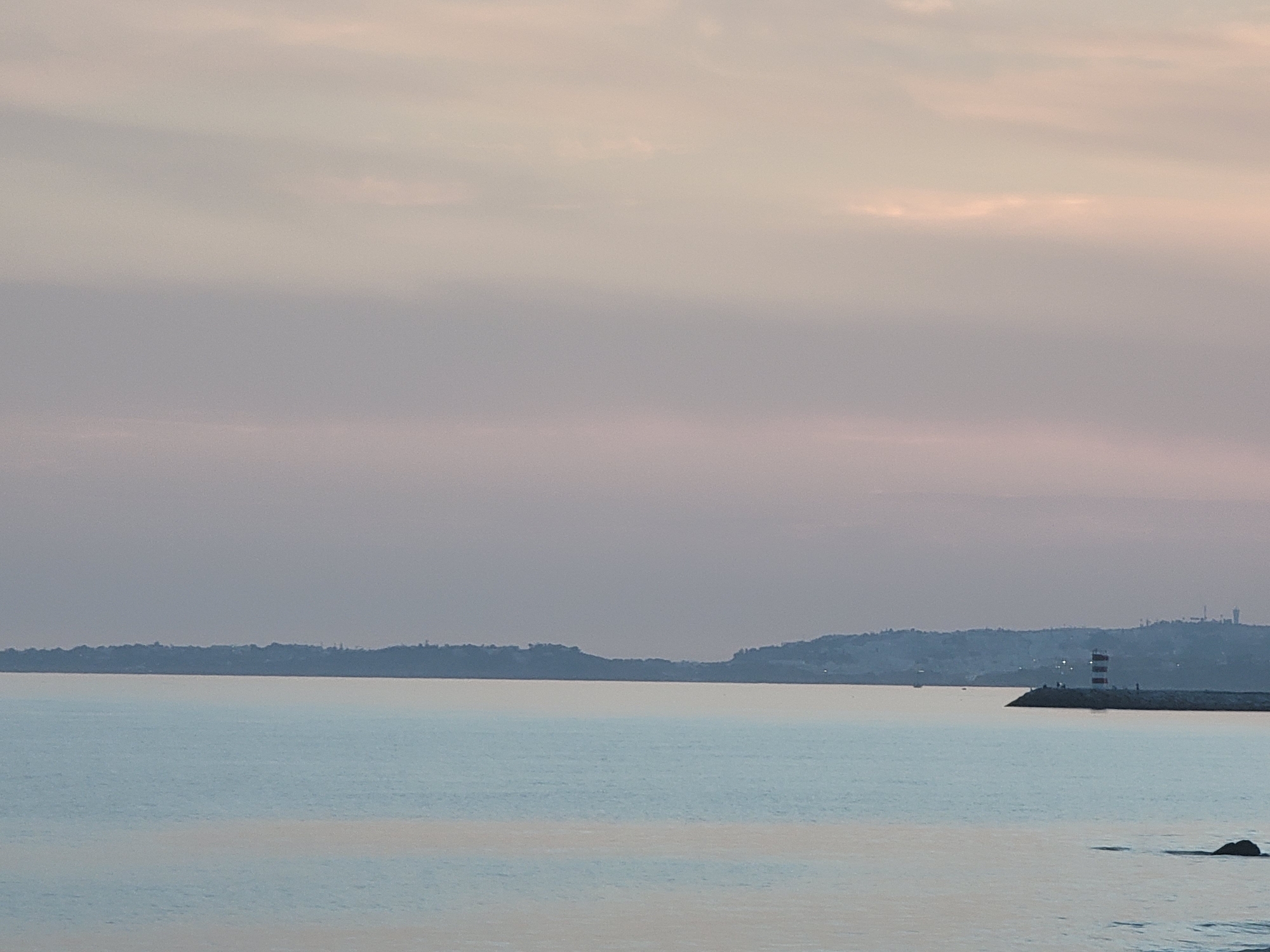 The image shows a body of calm water with a distant, hazy shoreline under a pale, overcast sky. On the right side of the image, a red and white striped lighthouse stands on a breakwater. Several figures are visible standing on the breakwater near the lighthouse. The water surface reflects the muted colors of the sky, creating a serene and somewhat melancholic atmosphere. In the distance, the outline of hills with scattered structures can be seen along the horizon. This scene is characteristic of the Quarteira waterfront in Portugal, likely captured during dawn or dusk.