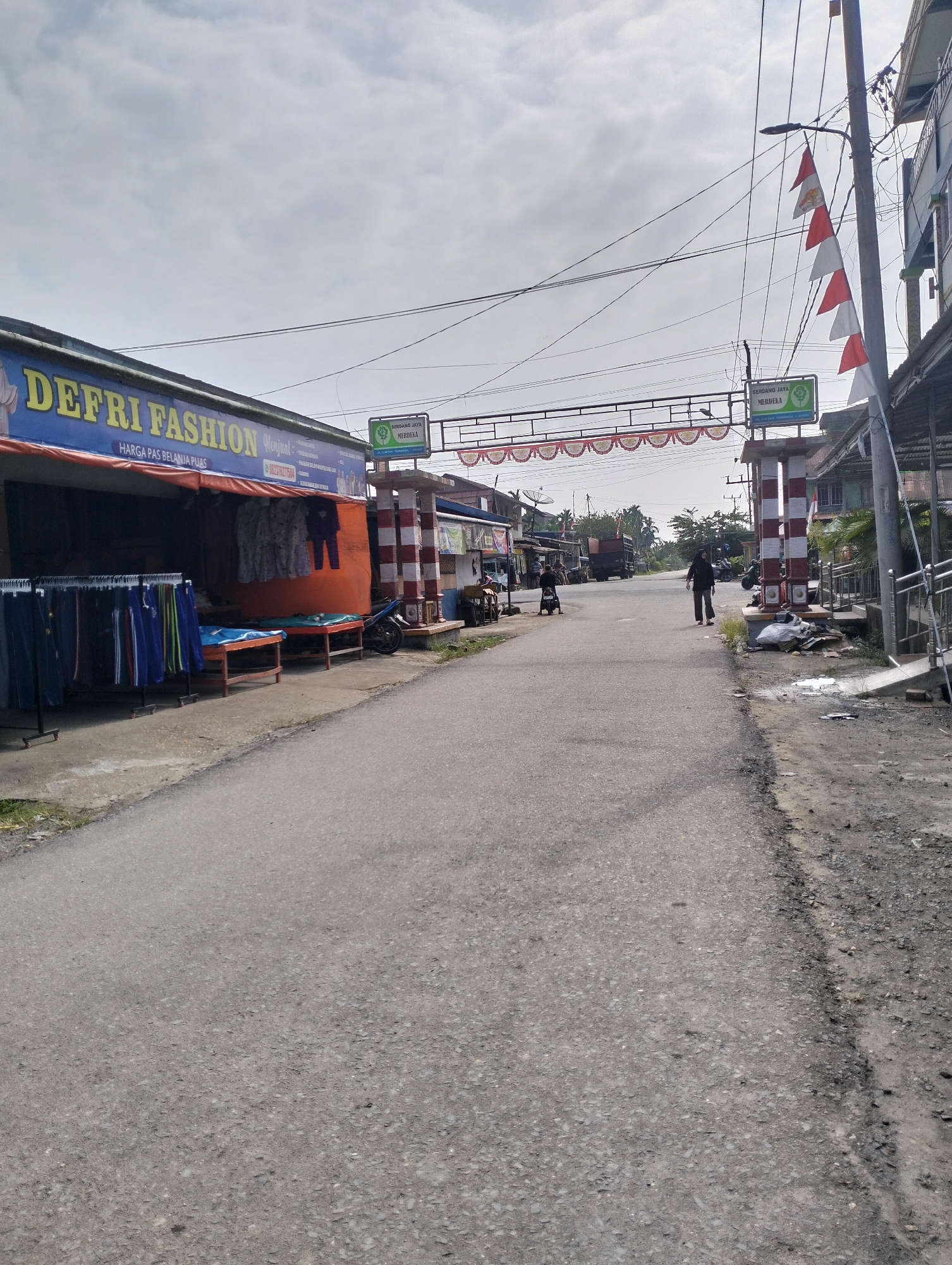 The image depicts a street scene in Pematang Lumut, Indonesia, likely during daylight hours under an overcast sky. On the left side of the road, a storefront identified as "DEFRI FASHION" is visible, displaying clothing on racks outside and through its open facade. The storefront has a blue awning with yellow lettering. Adjacent to the fashion store, a decorative archway spans the street, adorned with red and white pennant-like flags. A sign on a pole near the archway reads "SERDANG JAYA MERDEKA".
The road itself is paved and appears to be a two-lane street with a worn surface. In the middle ground, several people are present. One individual, a woman wearing a black headscarf and black clothing, is walking away from the camera down the center of the road. Other individuals are seen on motorcycles in the distance. Various vehicles, including a truck, are also present on the road. The right side of the street features buildings with tin roofs and a sidewalk area with a metal railing. Debris is visible on the side of the road near a striped pillar that forms part of the archway structure. The environment suggests a somewhat rural or semi-urban setting.
