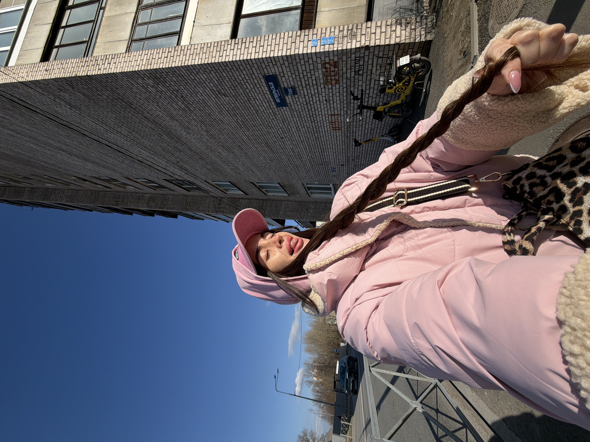 The image depicts a female individual in a selfie-style shot on an urban street in Saint Petersburg, Russia. The location is identified by a street sign on the adjacent building reading "ОКТЯБРЬСКАЯ" (Oktyabrskaya) and "10 корпус" (10 korpus).
The subject, positioned in the foreground, is wearing a light pink hooded puffer jacket with a cream sherpa lining visible at the cuffs and hood. A matching pink baseball cap is worn backward over the hood. She has long, dark hair styled in a single braid, which she holds with her right hand. Her face is angled slightly upwards with eyes closed and tongue partially extended, revealing freckles. A leopard-print shoulder bag with a black and white striped strap is visible on her left side. Her right hand displays manicured fingernails with a French tip design.
In the background, to the subject's right, is a multi-story residential building constructed of light grey brick and concrete panels, featuring multiple windows. At its base, several yellow electric scooters are parked, one with a visible license plate reading "УХ6 398". To the subject's left, a paved road is visible, separated by a grey metal pedestrian barrier. A dark-colored SUV is observed driving on the road. The far background includes bare trees and a street light pole under a clear blue sky with minimal cloud cover. The scene is illuminated by bright daylight, casting distinct shadows.