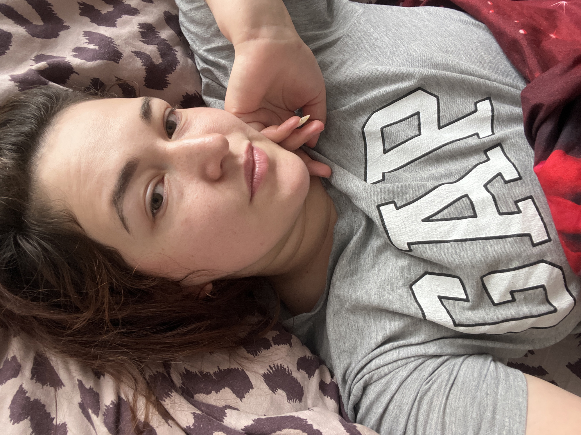 A young woman with dark hair is lying on a bed, resting her head on her hand. She is wearing a grey t-shirt with the "GAP" logo printed on it. The background shows a patterned comforter with a leopard-like print. The lighting is soft and natural. The scene is indoors. There are no specific details in the image that would allow for the determination of a location within the city of Chelyabinsk, Russia.