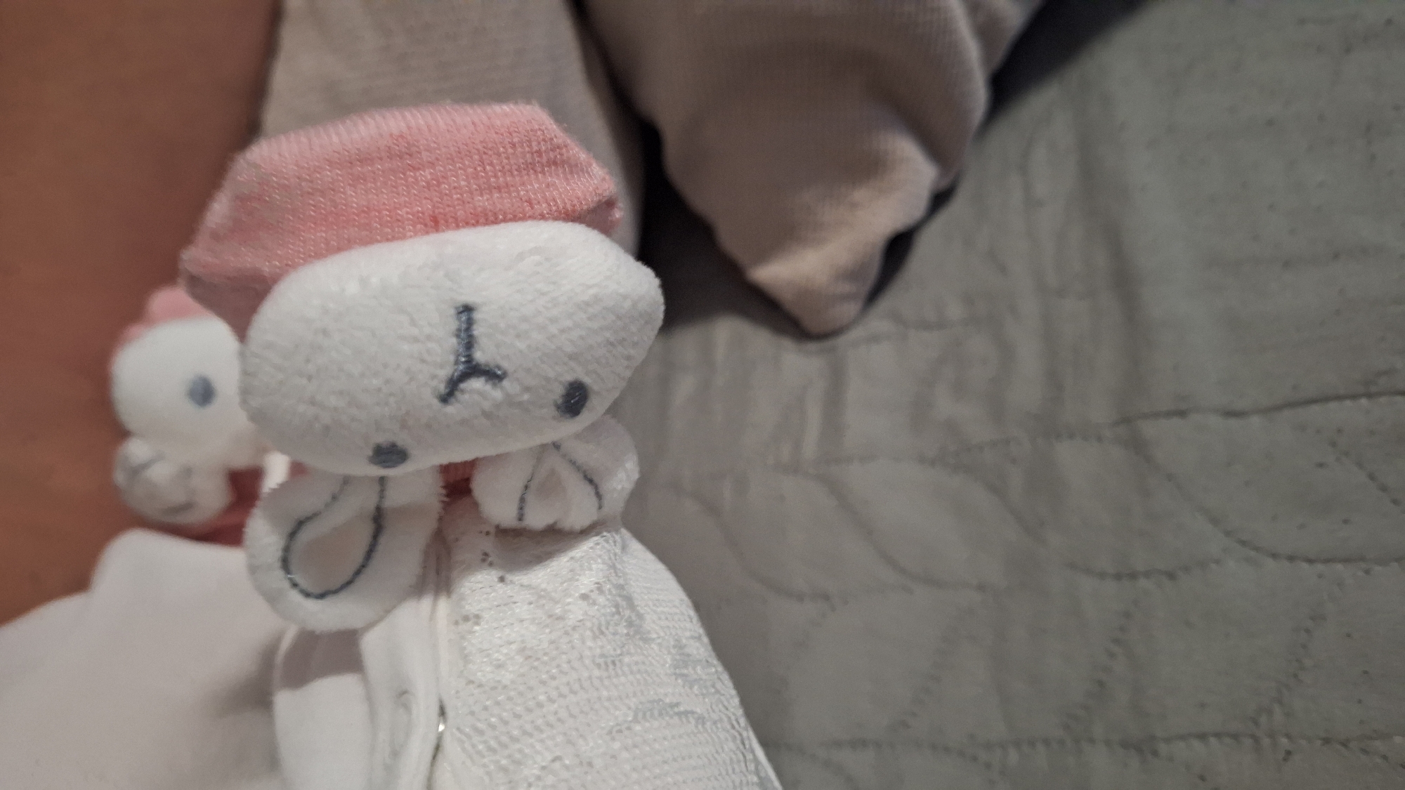 The image shows a close-up of a small, white plush toy with a pink hat and a gray stitched face. The toy appears to be a rabbit, with floppy ears and a white fabric body. Another similar plush toy is visible in the background, slightly out of focus. The toys are resting on a textured, gray surface that appears to be bedding. There is no information available within the image to ascertain the location in the city of Blagoevgrad, Bulgaria.
