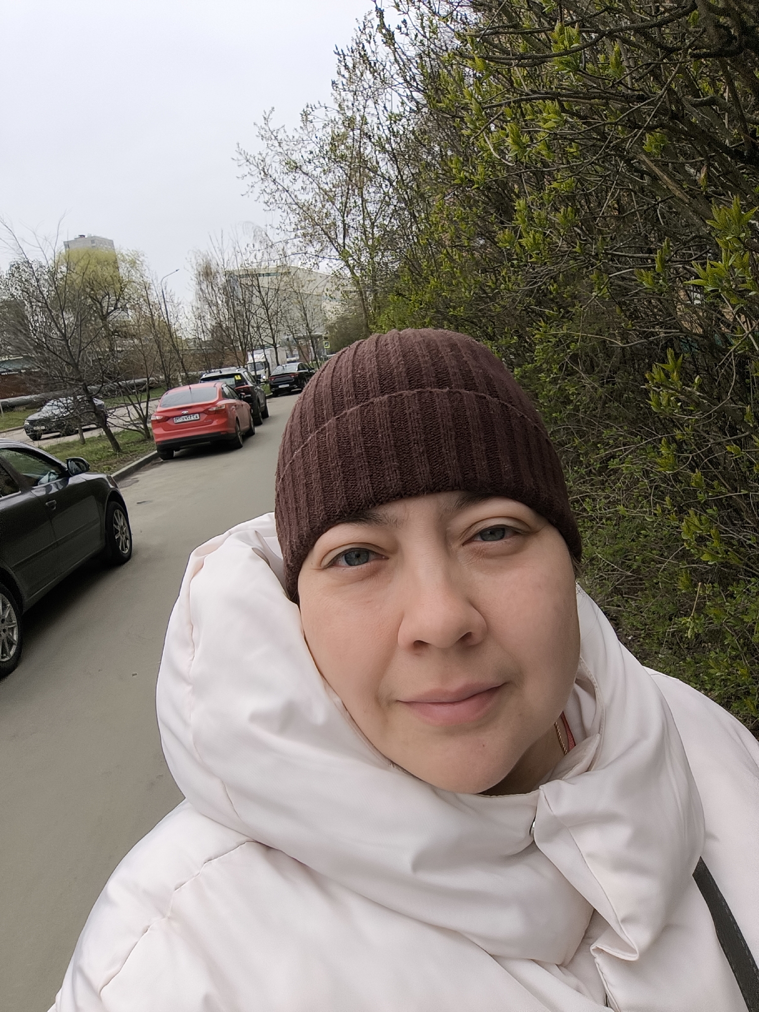 A selfie of a woman wearing a brown beanie and a light pink puffer jacket stands on a sidewalk in Khimki, Russia. The street behind her is lined with parked cars, and buildings are visible in the background. The trees are starting to show new leaves, suggesting it is spring.
