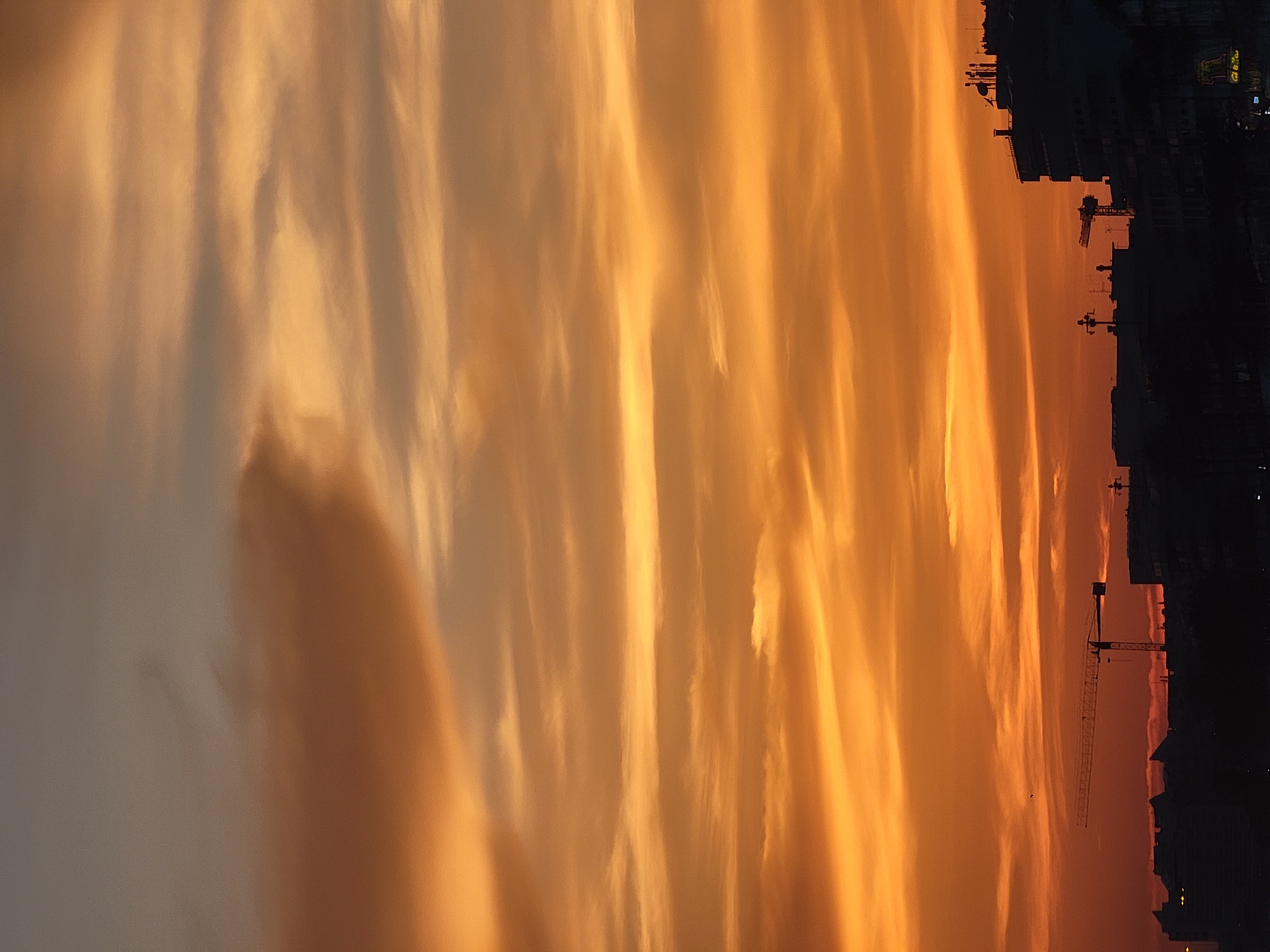 The image depicts a dramatic sunset over the city of Quarteira, Portugal. The sky is filled with streaks of orange, yellow, and brown clouds, creating a vibrant and textured backdrop. In the foreground, the silhouetted shapes of buildings and a construction crane are visible against the colorful sky. The overall scene is one of a developing urban landscape set against a beautiful natural phenomenon.