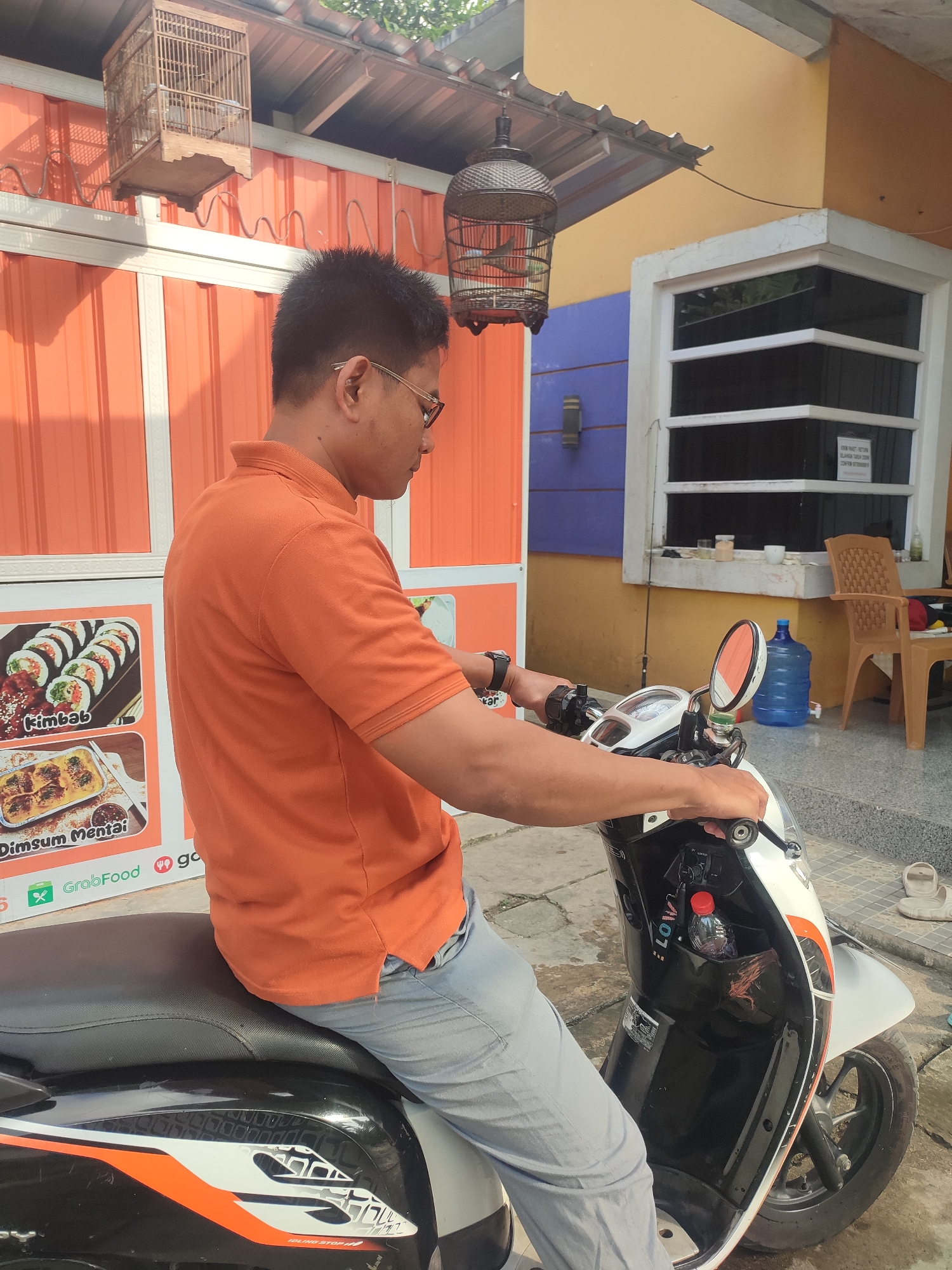 A medium shot shows a man wearing an orange polo shirt and light gray pants, seated on a white and black motorcycle with orange accents. He is looking to his right, with his hands on the handlebars. The man has short dark hair and wears glasses. In the background, there is an orange corrugated metal wall with a white trim. Above the wall, a birdcage hangs from a corrugated roof. A second, darker birdcage is partially visible on the right side of the frame. Further to the right, a yellow wall features a window with dark panes and white trim. Adjacent to the window is a brown plastic chair. A large blue water jug is positioned near the window. The man is positioned in front of a display featuring images of food, including kimbab and dimsum mentai, with a "GrabFood" logo visible. The ground is paved with gray tiles. The scene appears to be in Sarwadadi, Indonesia.