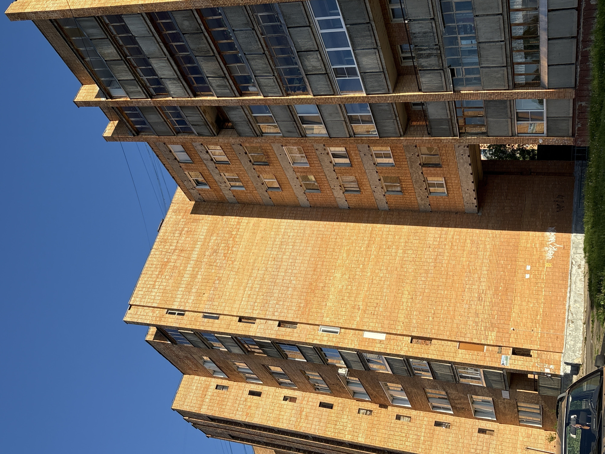 This is an exterior, low-angle, vertical-format, medium shot of an old, multi-story brick apartment building under a clear blue sky. The building is constructed of orange-brown bricks and features numerous balconies with gray, weathered railings. Many of the windows are visible, some reflecting the sunlight. A passage or archway is present between sections of the building, revealing a glimpse of trees and foliage beyond. The building appears to be located in a residential area, with a patch of grass and a portion of a car visible in the foreground. There are no people in the frame. The building's architecture suggests it is likely from the Soviet era, typical of many buildings found in cities like Kirovo-Chepetsk, Russia.