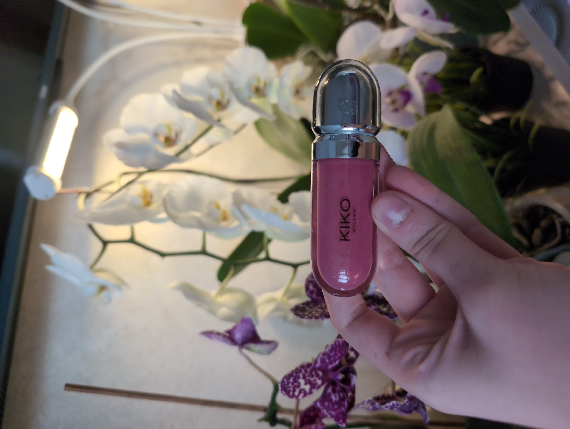 The scene features a hand holding a KIKO Milano lip gloss tube against a backdrop of white and purple orchids. A small LED light is visible in the background, suggesting indoor lighting. The location is not specified, and no clues in the image suggest its city or country.